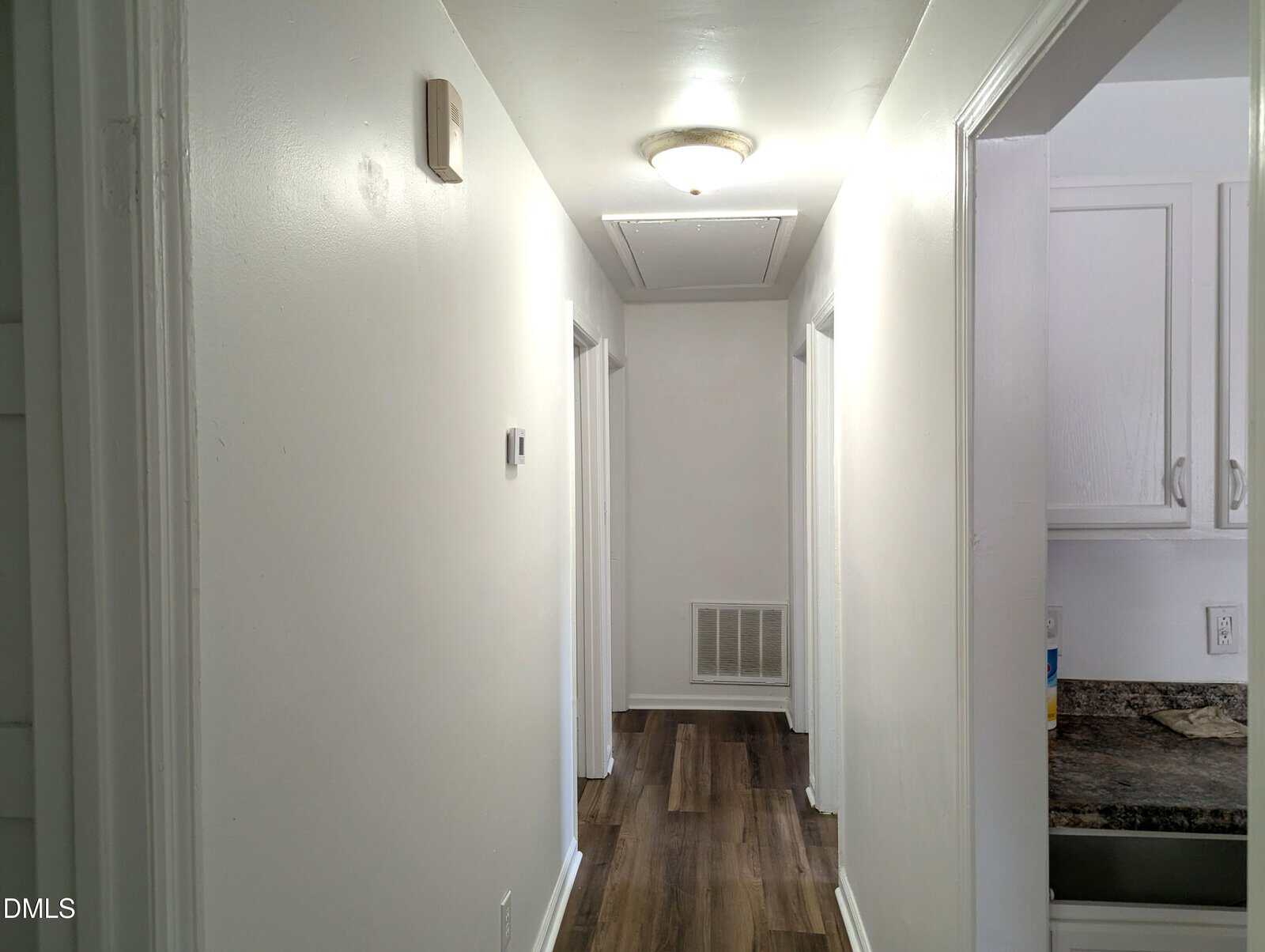 A brightly lit hallway with white walls and ceiling, wood-style flooring, and several doorways; a kitchen with white cabinets and dark countertops is partially visible on the right.