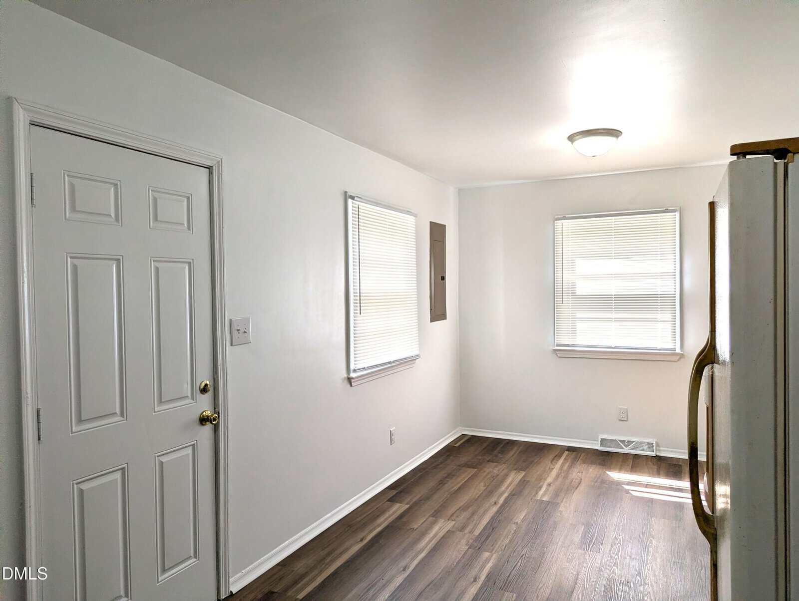 Bright kitchen with white walls, two windows with blinds, a door, and wood-style flooring. A refrigerator is partially visible on the right side of the image. Ceiling light fixture is turned on.