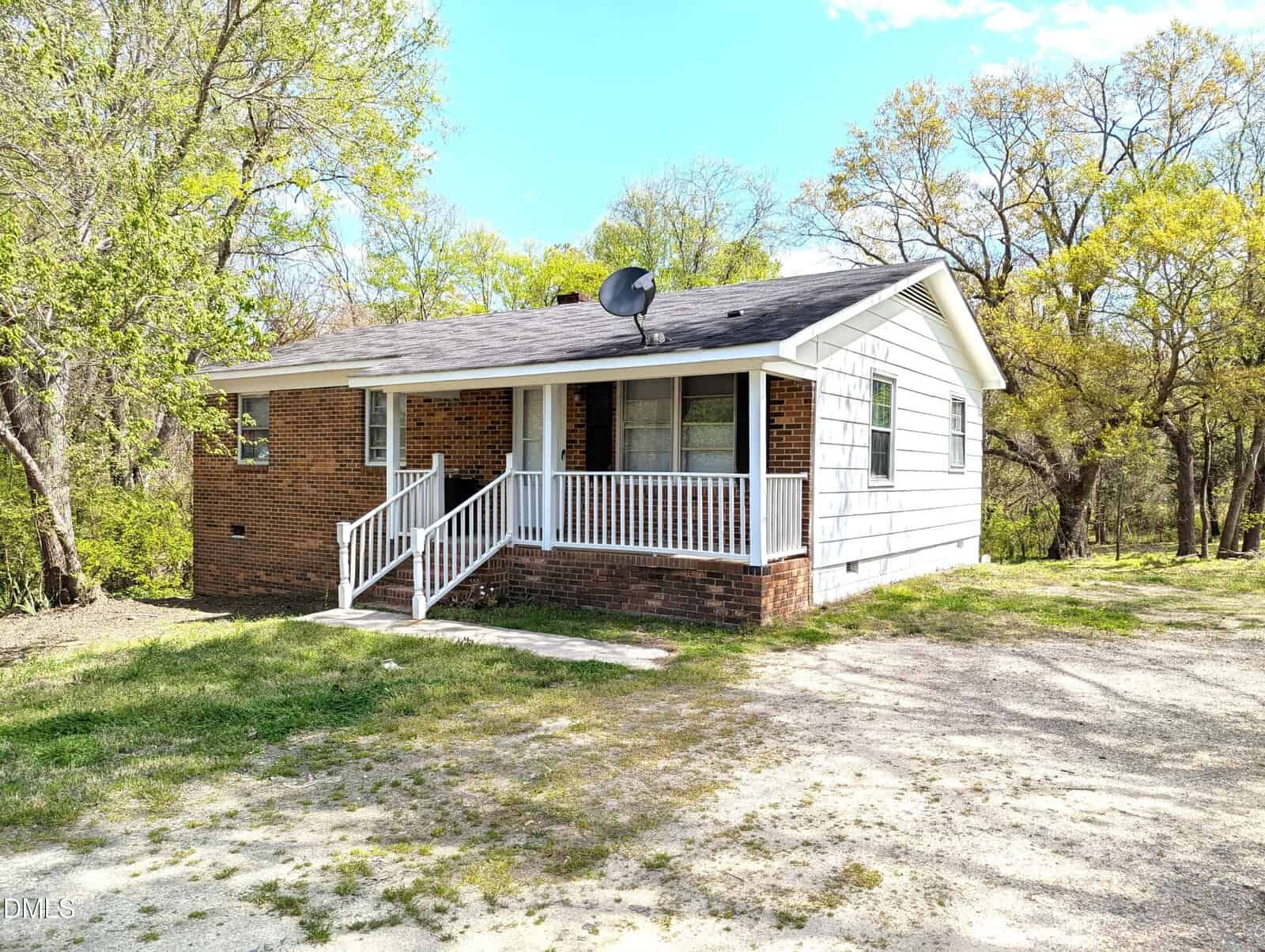 A small, single-story brick and white-sided house with a front porch, white railing, and satellite dish on the roof, surrounded by trees and a grassy yard.
