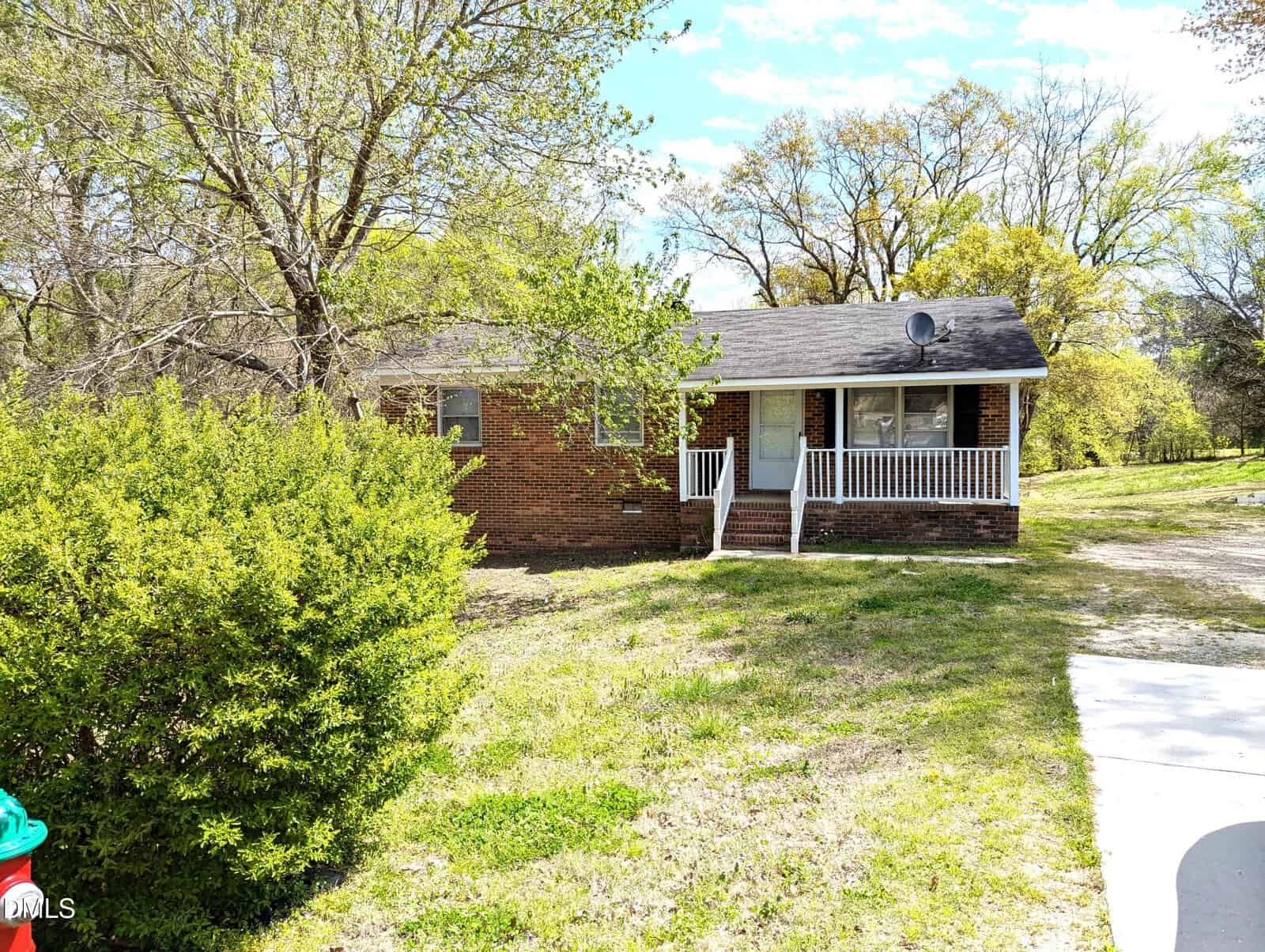 A single-story brick house with a small front porch, white railing, and steps leading to the entrance. The yard has sparse grass and mature trees, with a concrete driveway visible on the right.