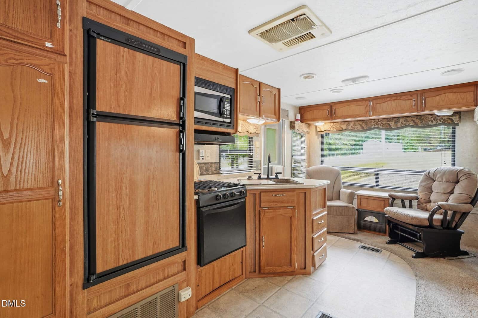 Interior of an RV with wooden cabinets, a small kitchen area featuring a stove, oven, microwave, and fridge. There are two cushioned chairs near large windows with blinds, and natural light fills the space.