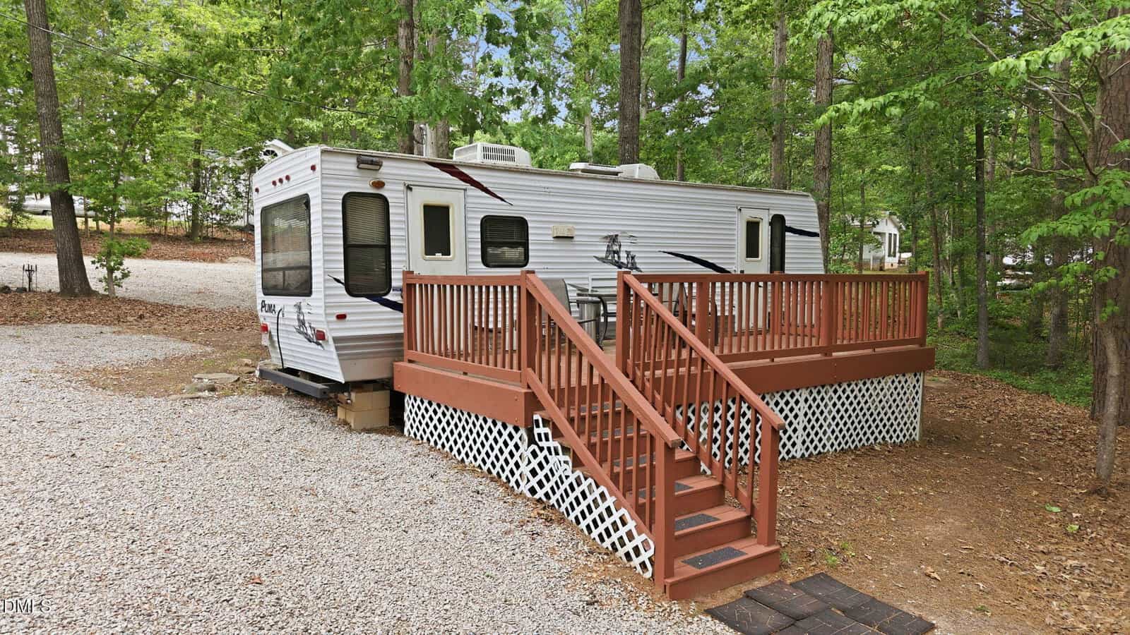 A white travel trailer with maroon and black accents sits in a wooded area, next to a wooden deck with railings and lattice skirting. Gravel surrounds the trailer, and tall green trees provide shade.