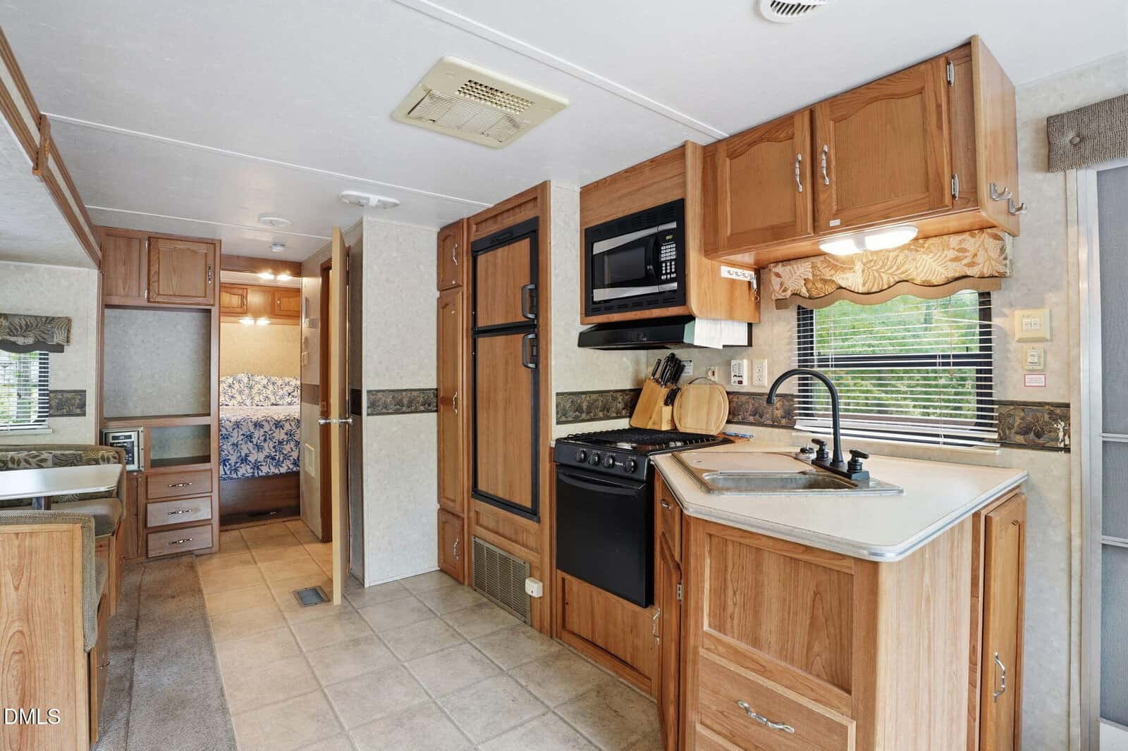 Interior of an RV kitchen with wood cabinets, a stove, oven, microwave, sink, and a window with blinds. A hallway leads to a bedroom with a blue-patterned bedspread. The floor is tiled, and counters hold kitchen essentials.