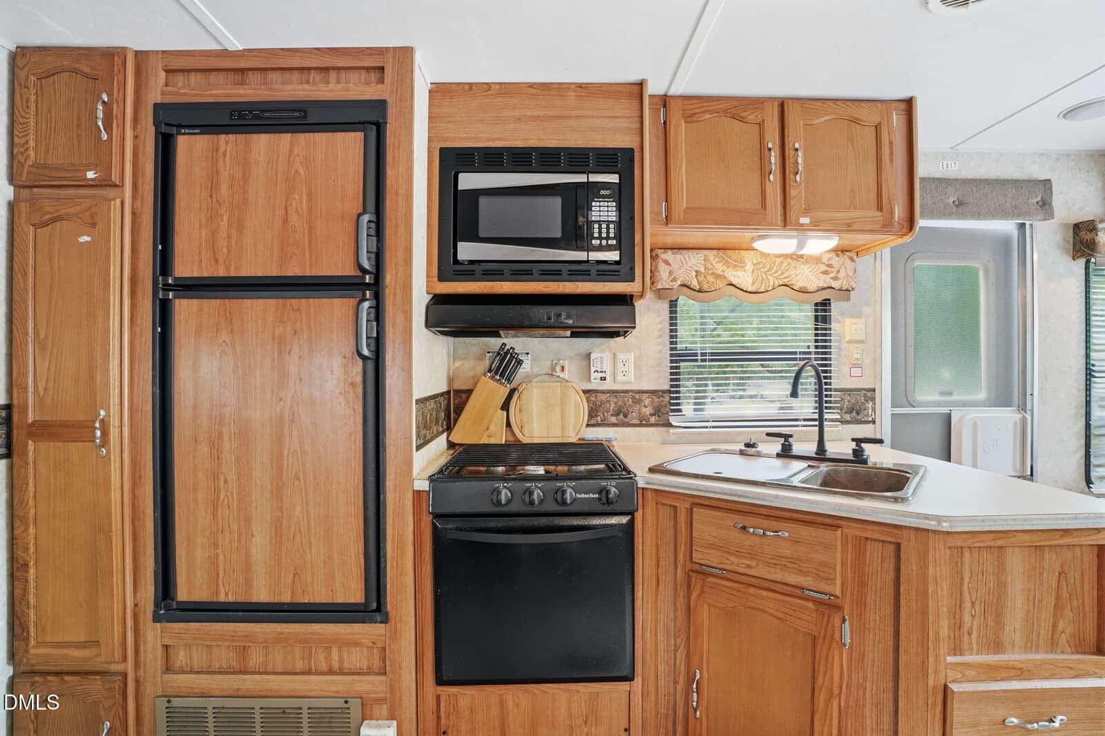 Kitchen area in an RV with a wood-paneled fridge, black stove and microwave, sink, cabinets, and a window with a valance. Bright natural light streams through the window and door.