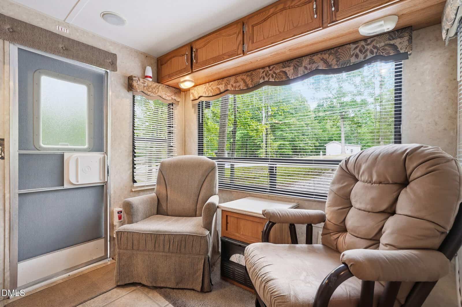 Two cushioned chairs sit next to a small table in an RV, with large windows behind them revealing a view of green trees and an outdoor scene. Overhead cabinets and a door are also visible in the cozy space.