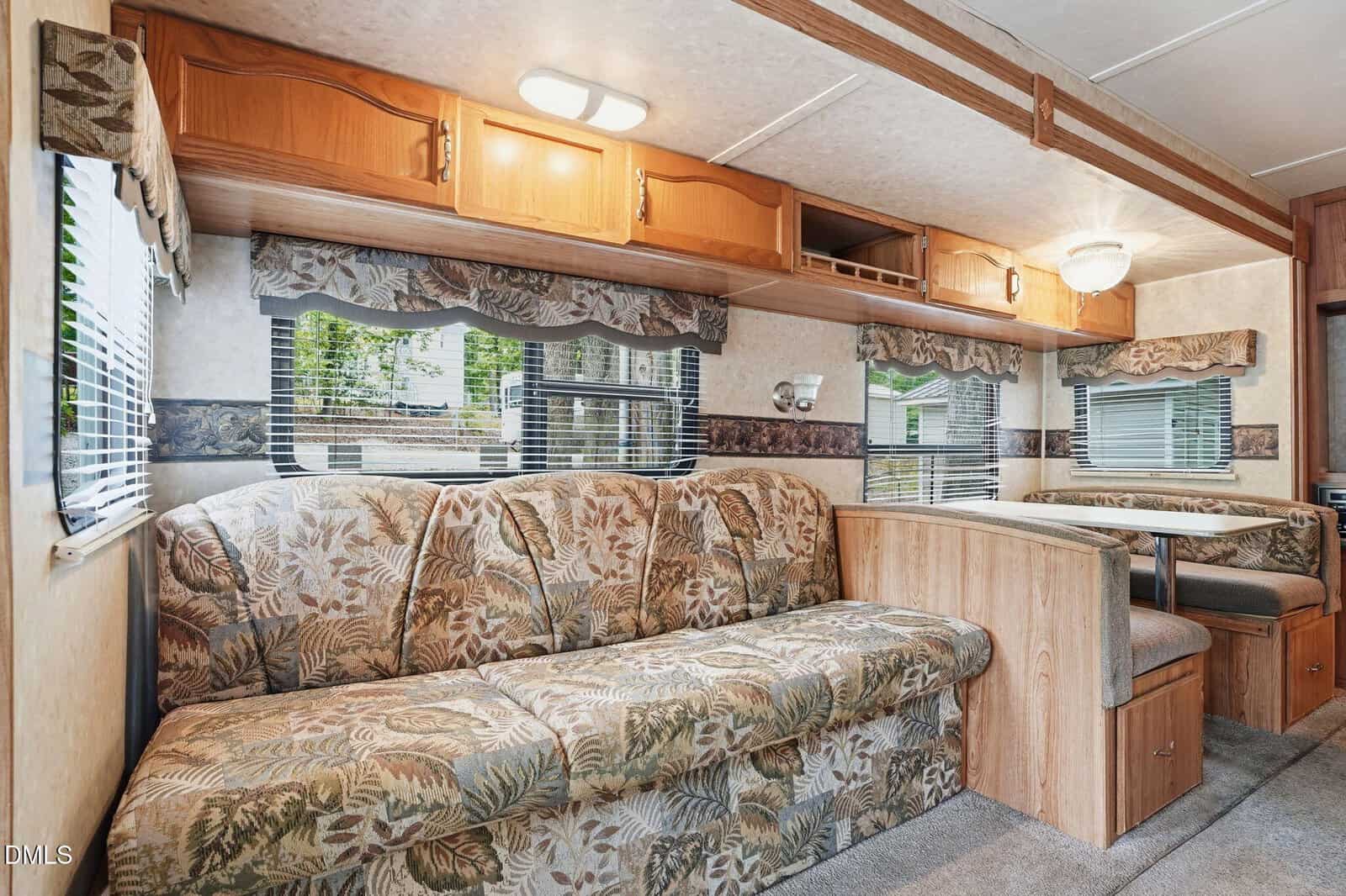 Interior of an RV with a floral-patterned sofa, wooden cabinets above, and a dining booth with seats. Multiple windows let in natural light, creating a cozy and inviting space.
