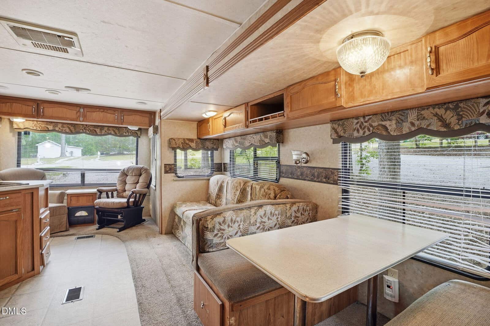 The interior of an RV features a dining table with bench seats, a floral-patterned sofa, a wooden chair, and large windows with views outside. Light wood cabinets and beige decor create a cozy, inviting atmosphere.