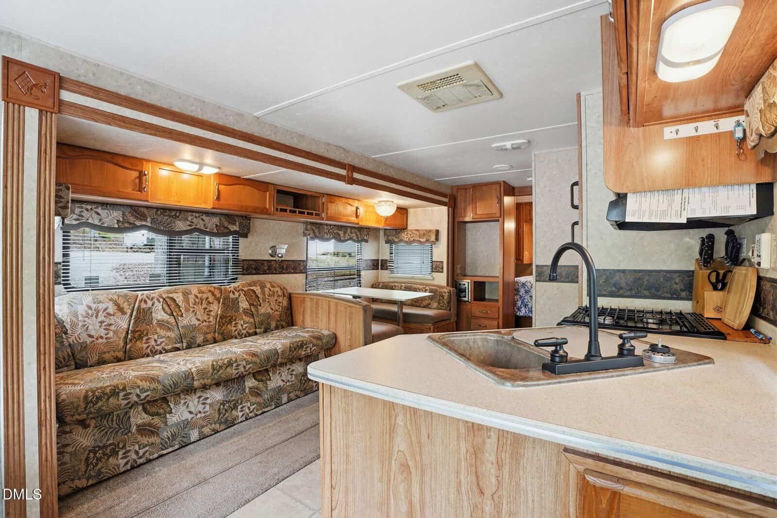 Interior of an RV featuring a floral-patterned sofa, dining table, kitchen counter with sink and faucet, stove, wooden cabinets, large windows with blinds, and a hallway leading to another area.