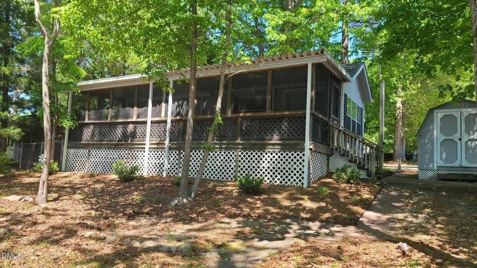A single-story house with a large screened porch and lattice skirting, surrounded by trees. A gray shed sits to the right along a concrete path, and sunlight filters through the leafy canopy above.