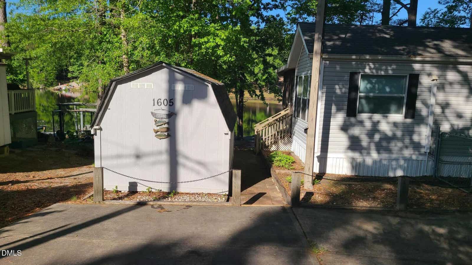 Small white shed with the number 1605 on the door, situated between a beige mobile home and a driveway, surrounded by trees, with a glimpse of a lake in the background.