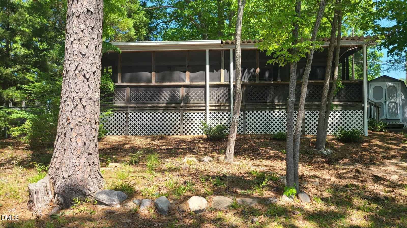 A screened-in porch with a lattice foundation is surrounded by trees and shaded by sunlight. The ground is covered with mulch, rocks, and sparse plants. A small shed is visible in the background.
