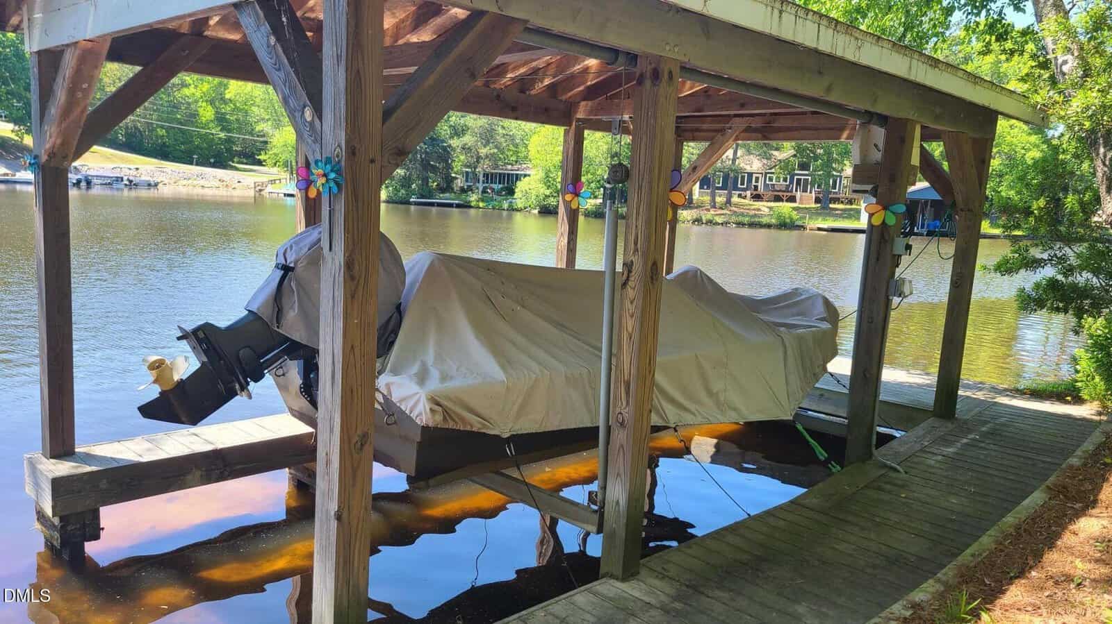 A covered wooden boat dock holds two boats with protective covers next to a calm lake, surrounded by trees and houses. Colorful pinwheels decorate the dock beams. Sunlight filters through the leaves.