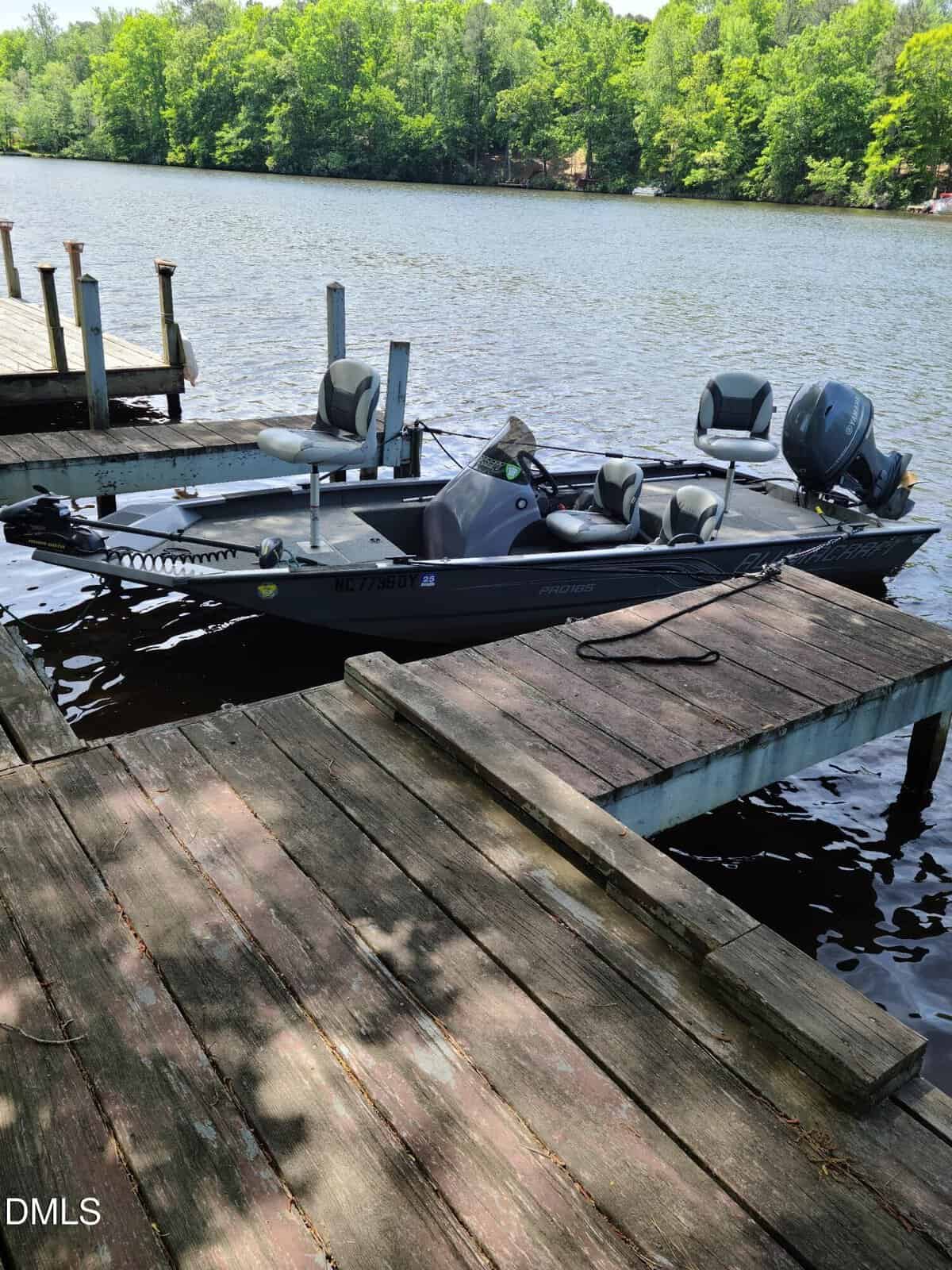 A small fishing boat is docked at a wooden pier on a calm lake, surrounded by green trees under a bright sky. The water is still and the area looks peaceful and quiet.