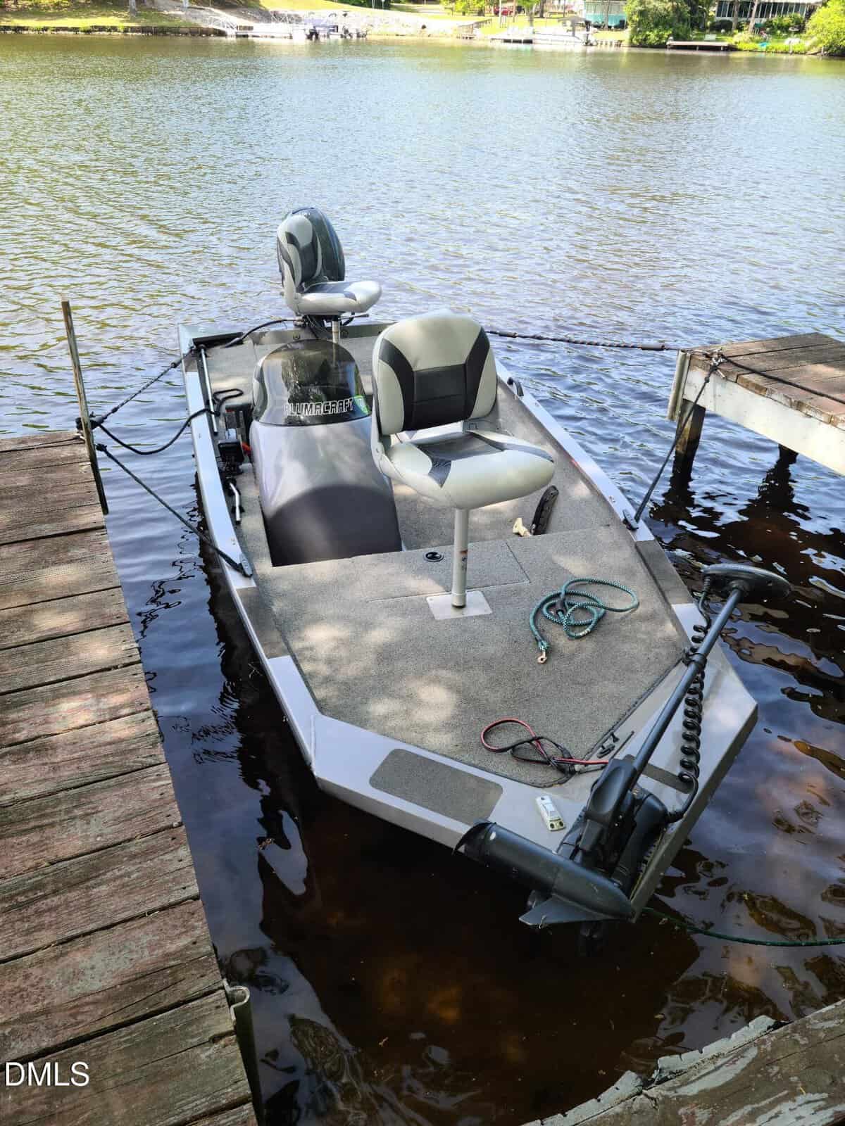 A small fishing boat with two seats is docked at a wooden pier on a calm lake. Some fishing equipment and cables are visible on the boat’s deck. Trees and homes are visible across the water.