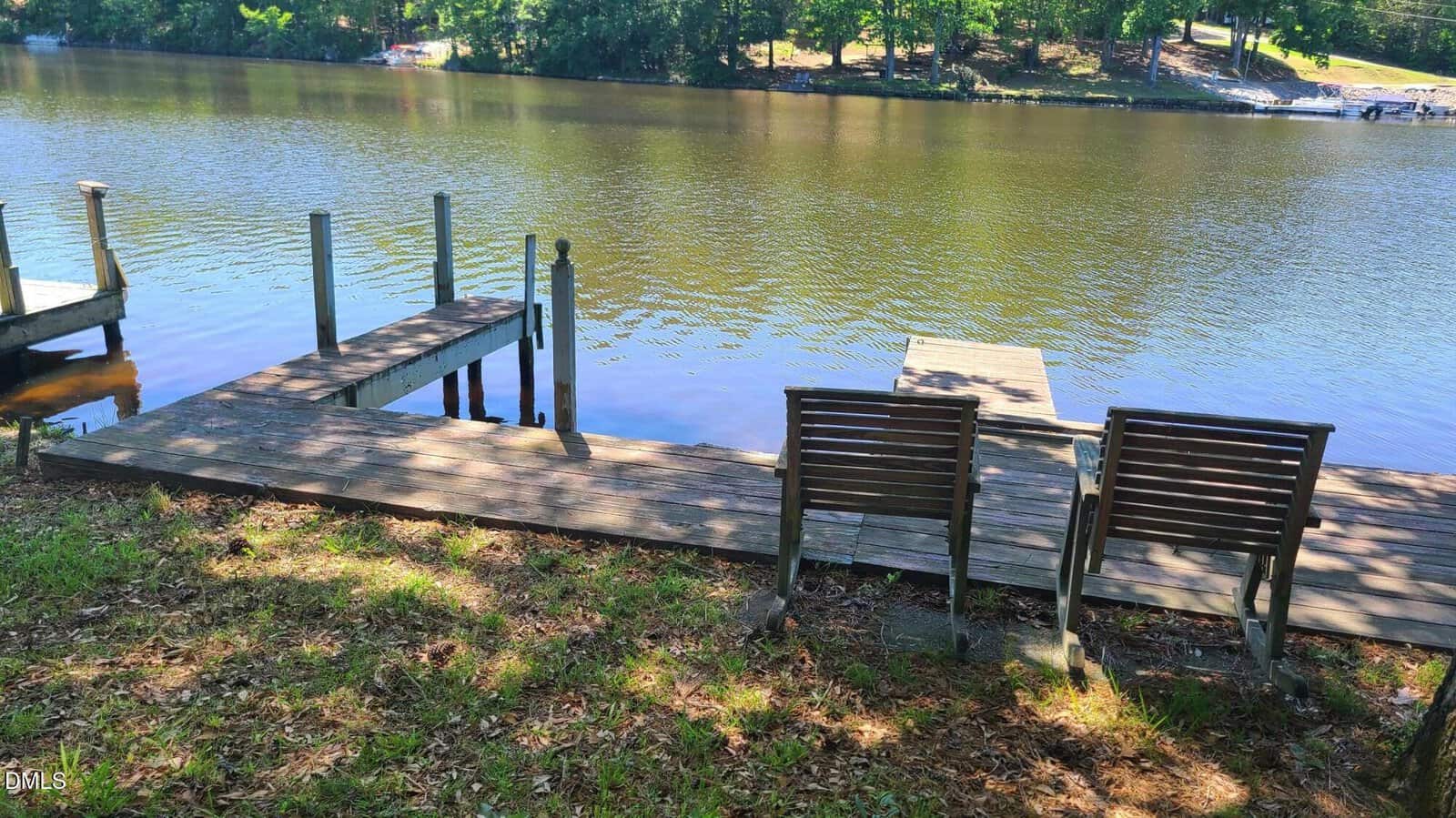 Two wooden chairs face a calm river, positioned beside a wooden dock. Trees and greenery line the opposite riverbank under a bright, sunny sky.