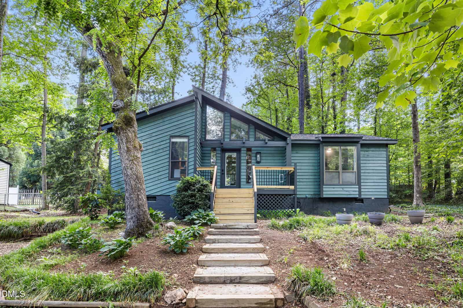 A modern blue house with large windows and a small front porch, surrounded by tall green trees and a landscaped yard with stone steps leading to the entrance.