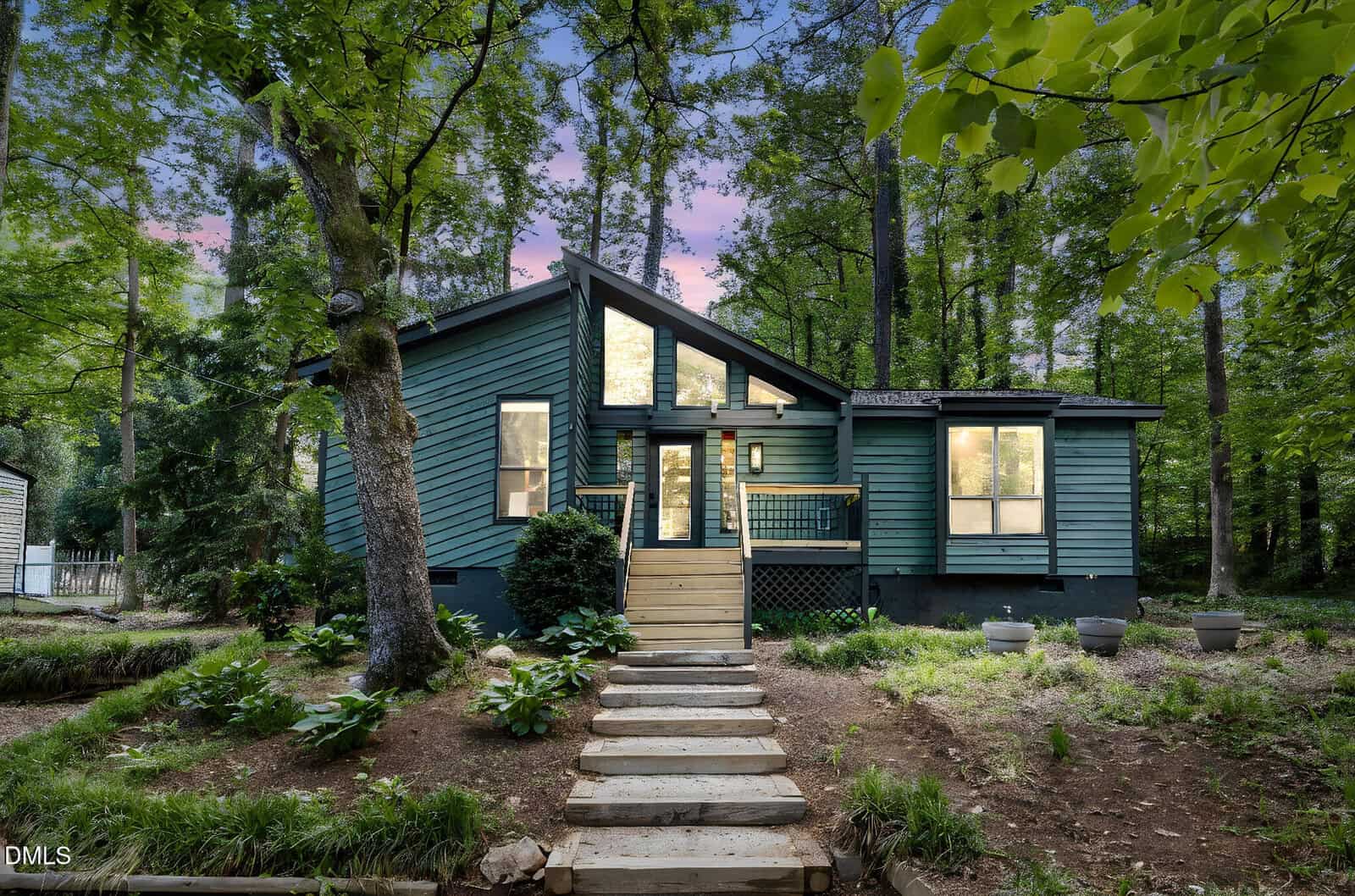 A modern blue house with large windows and a front porch, surrounded by tall green trees. Stone steps lead up to the entrance, and the setting is lush and wooded, with a pink and purple sunset sky in the background.