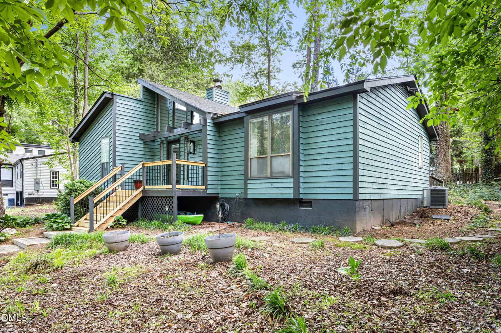 A light blue house with wood accents and a small back porch sits among tall trees, surrounded by a mulched yard with potted plants and stepping stones leading up to the stairs.
