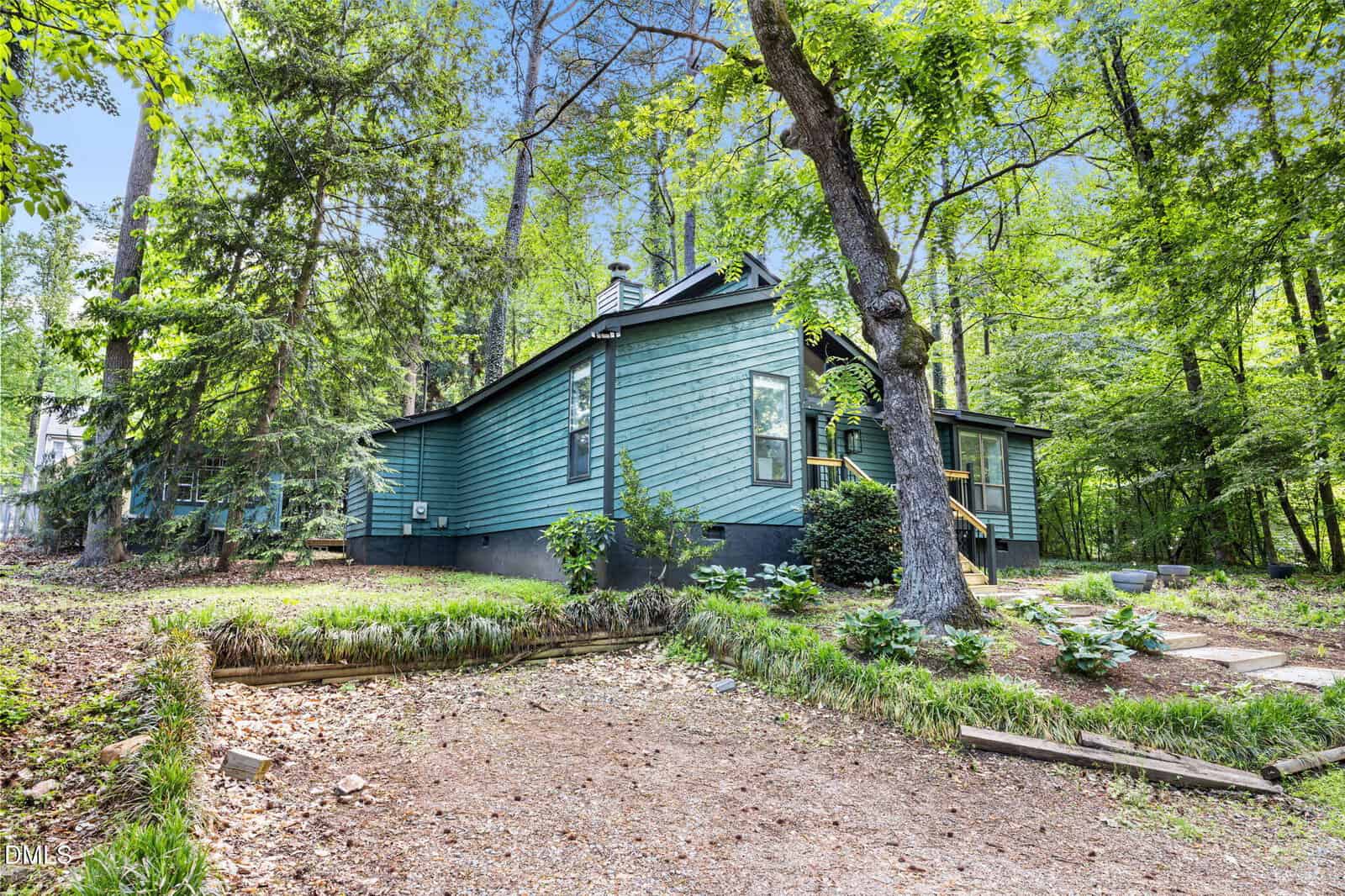 A green house with black trim sits among tall trees and lush greenery, with a gravel driveway and wooden steps leading to the front door. The surrounding yard is shaded and has patches of grass and garden beds.