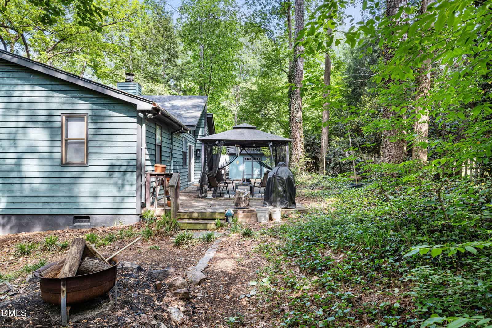 A green wooden house with a backyard patio featuring a gazebo, grill, outdoor furniture, and a fire pit surrounded by trees and lush greenery.