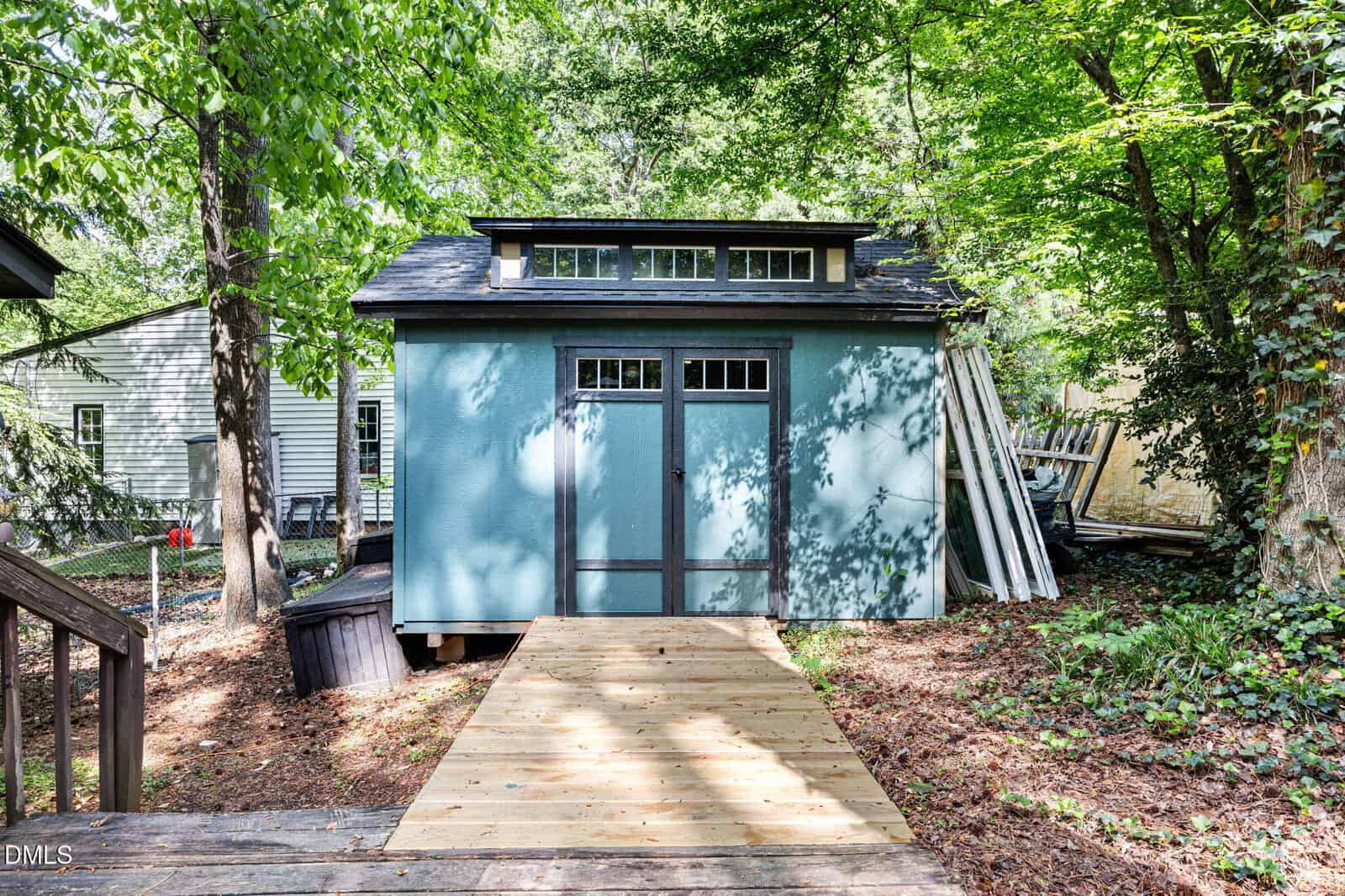A blue shed with double doors and transom windows sits in a wooded backyard, with a wooden ramp leading up to the entrance. Tall trees and neighboring houses surround the area.