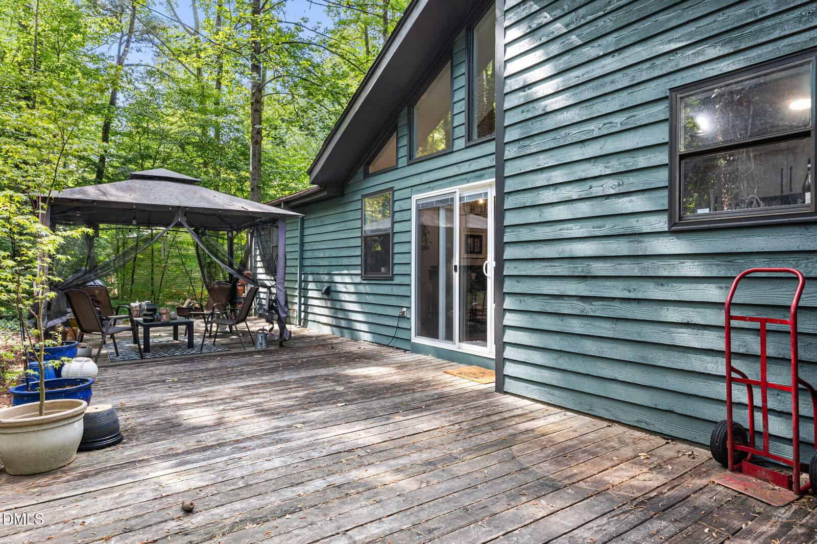 A wooden deck with potted plants, a red dolly, patio furniture under a canopy, and sliding glass doors. The house exterior is green with black-trimmed windows, surrounded by trees.