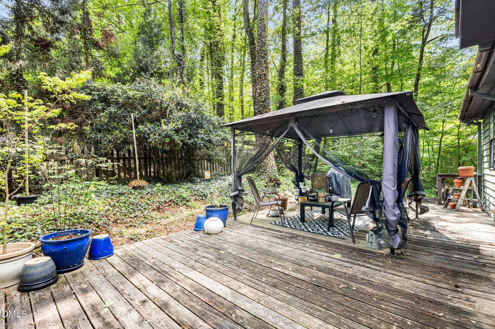 Wooden deck in a backyard with a canopy tent, outdoor furniture, potted plants, and surrounded by lush green trees and a wooden fence.