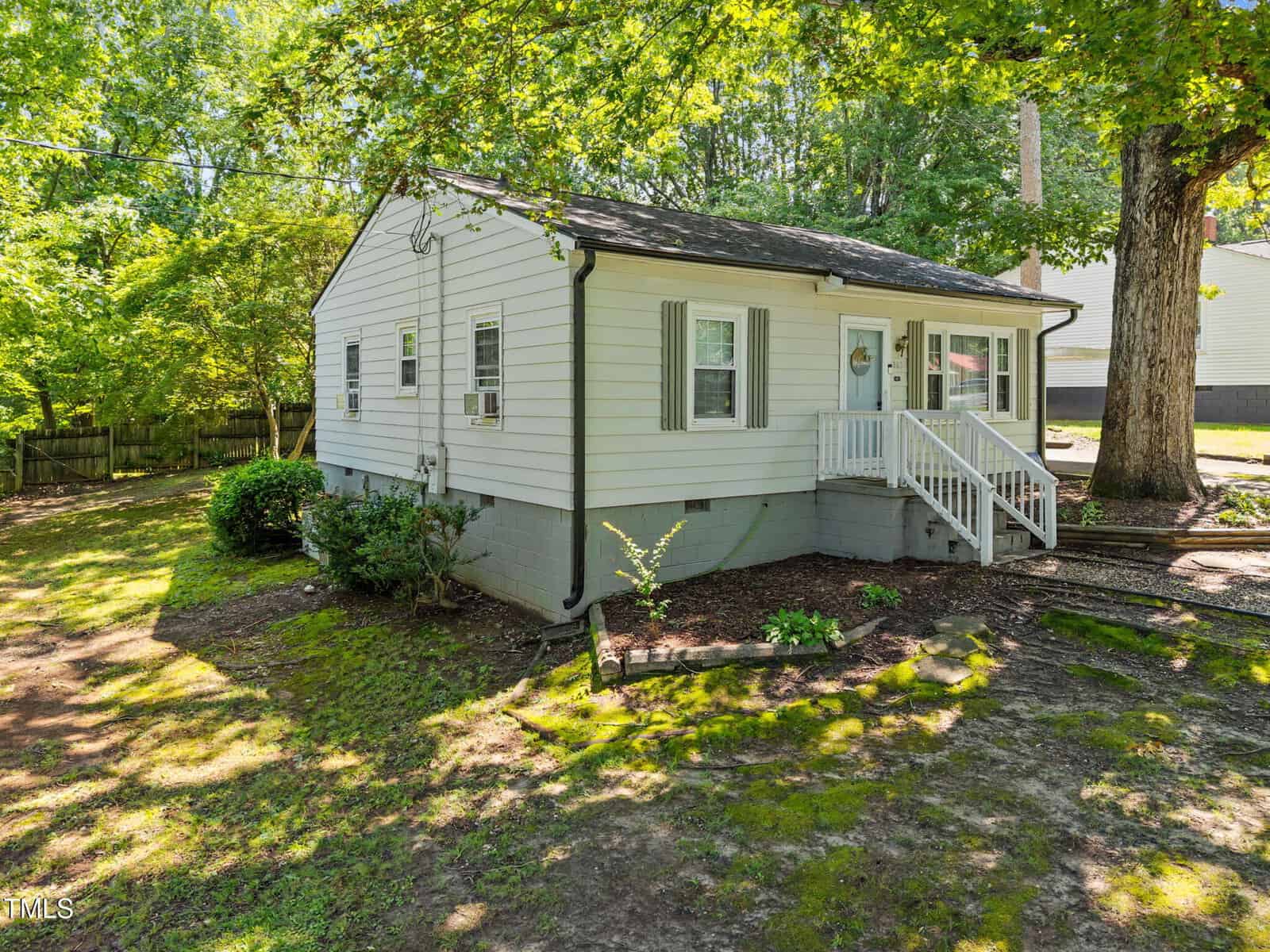 A small white one-story house with a front porch, green shutters, and a sloped yard with trees and bushes, surrounded by greenery and partially shaded by large trees.