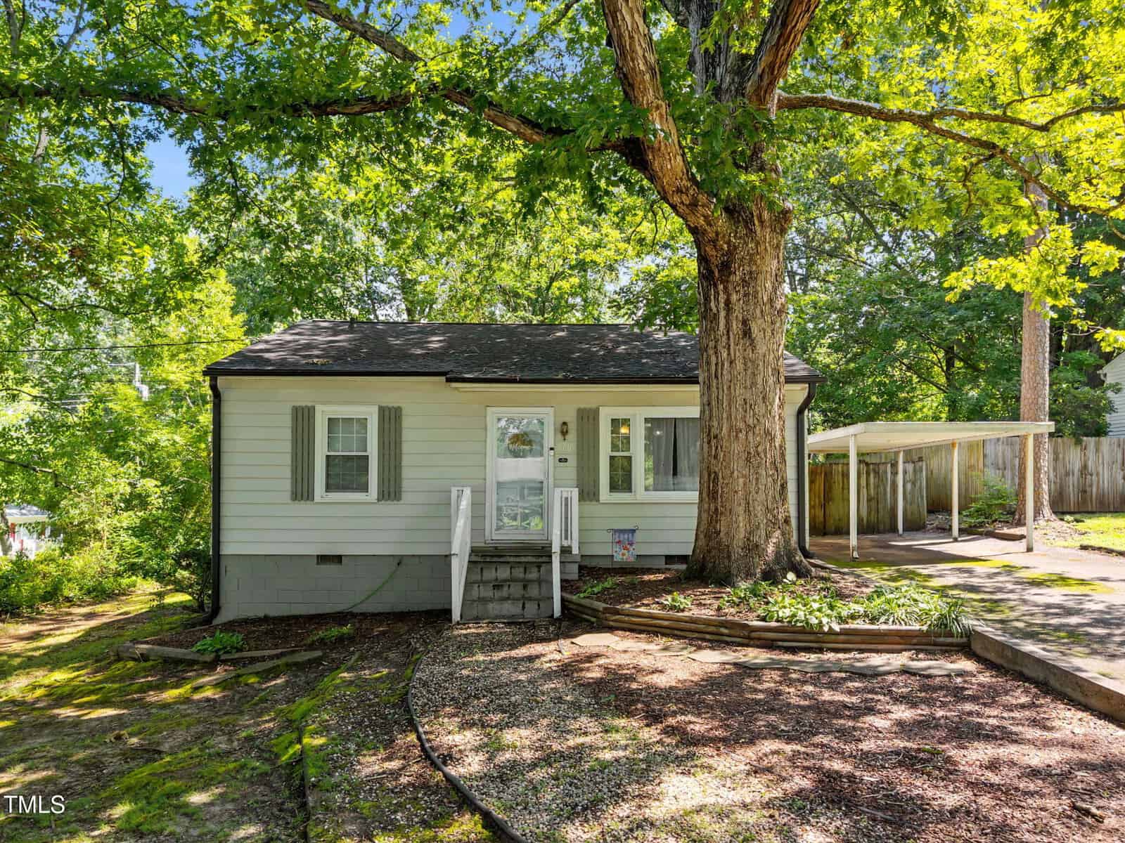 A small, light green house with white trim and shutters sits beneath a large tree. A short porch with steps leads to the front door, and a driveway with a carport is on the right. The yard is shaded and landscaped with mulch beds.