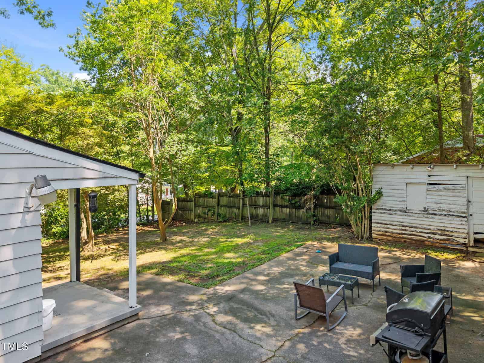 A backyard with a concrete patio featuring outdoor seating, a fire pit, and a grill. The yard is surrounded by trees and a wooden fence, with a small white shed in the background and a covered porch on the left.