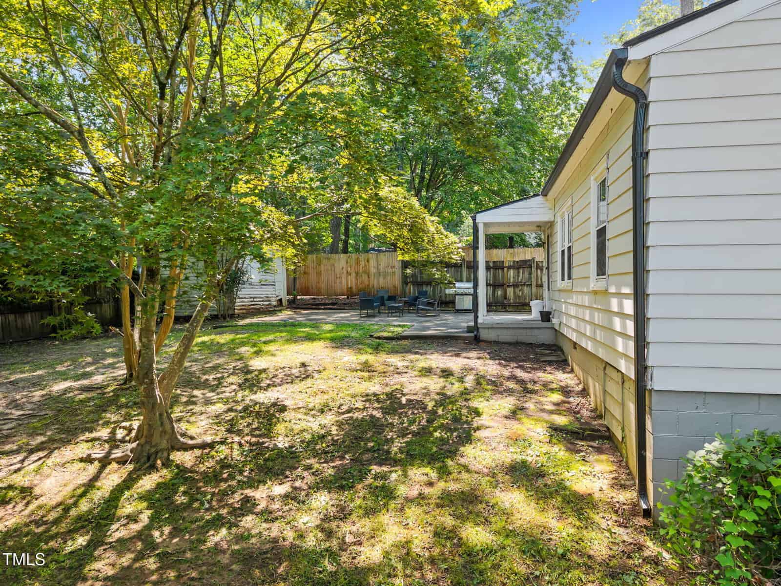 A sunny backyard with a tree, grassy lawn, wooden fence, patio area with seating, and a white house with horizontal siding on the right.