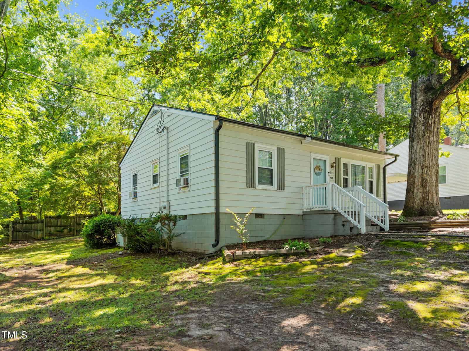 Single-story white house with green shutters, small front porch, and steps. Large trees provide shade, and there is a patchy lawn with some shrubs along the side.