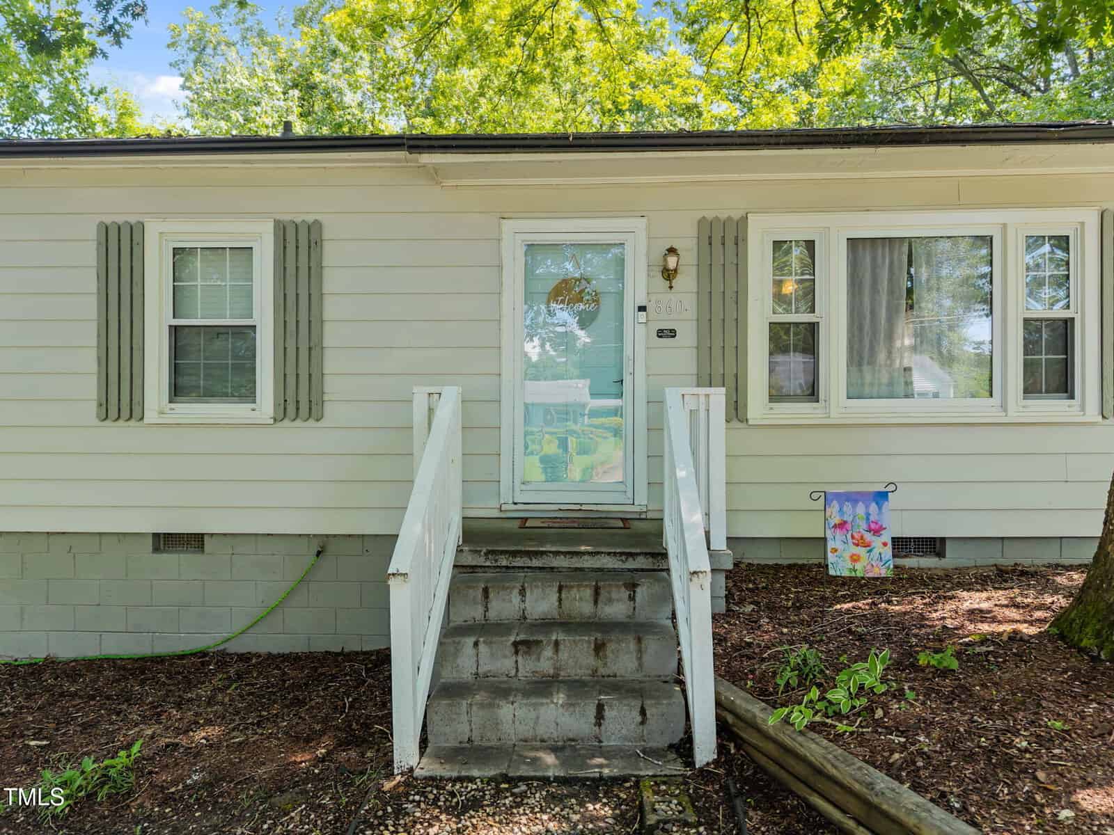Small, light-colored house with a glass storm door, steps leading to the entrance, gray shutters, multiple windows, and a colorful garden flag. Trees and mulch are visible in the foreground.
