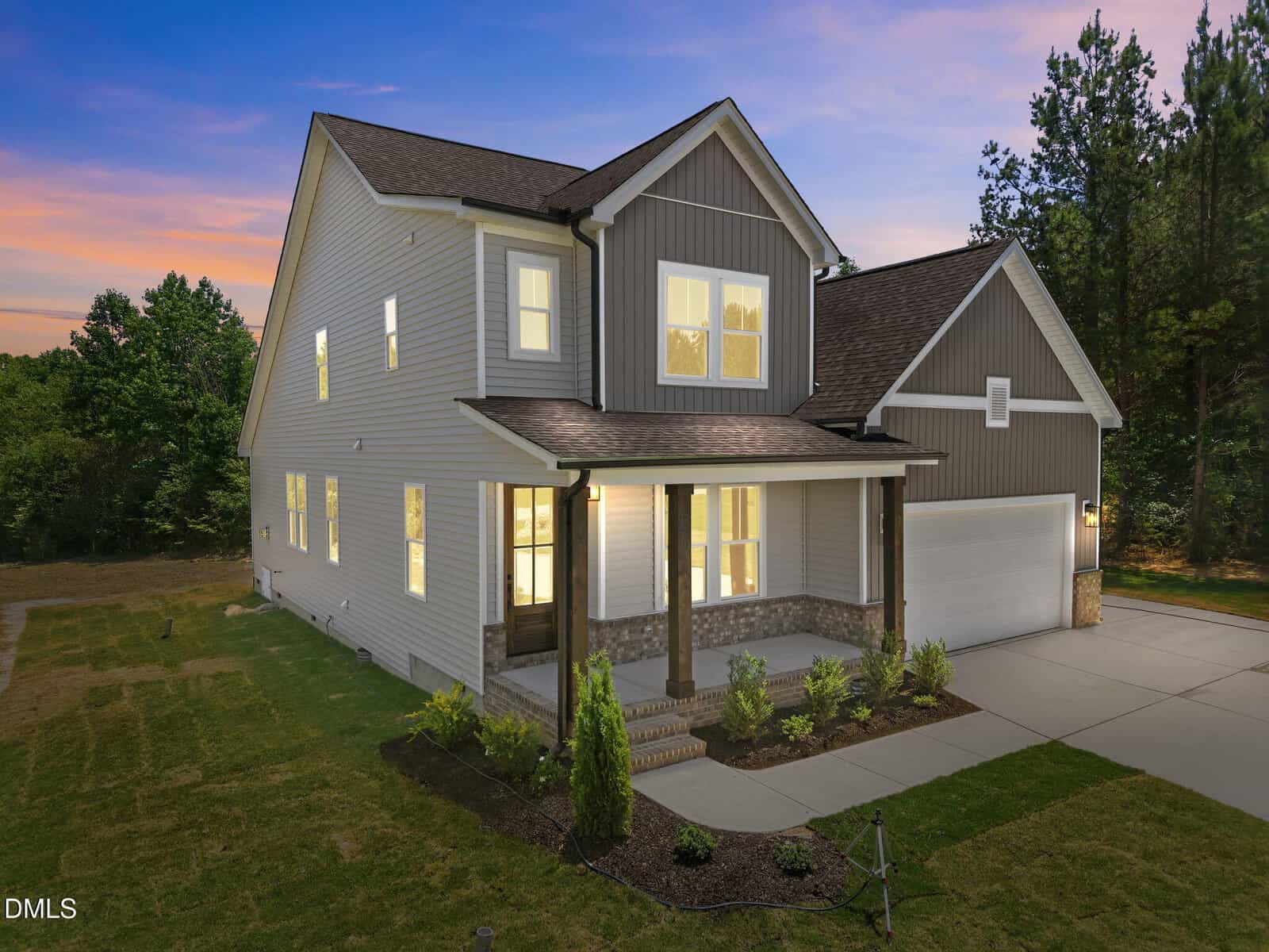Two-story modern suburban house at 80 Basil Court, Franklinton, with gray siding, a columned front porch, stone accents, double garage, landscaped yard, and exterior lights aglow at dusk. Surrounded by trees beneath a sunset sky.