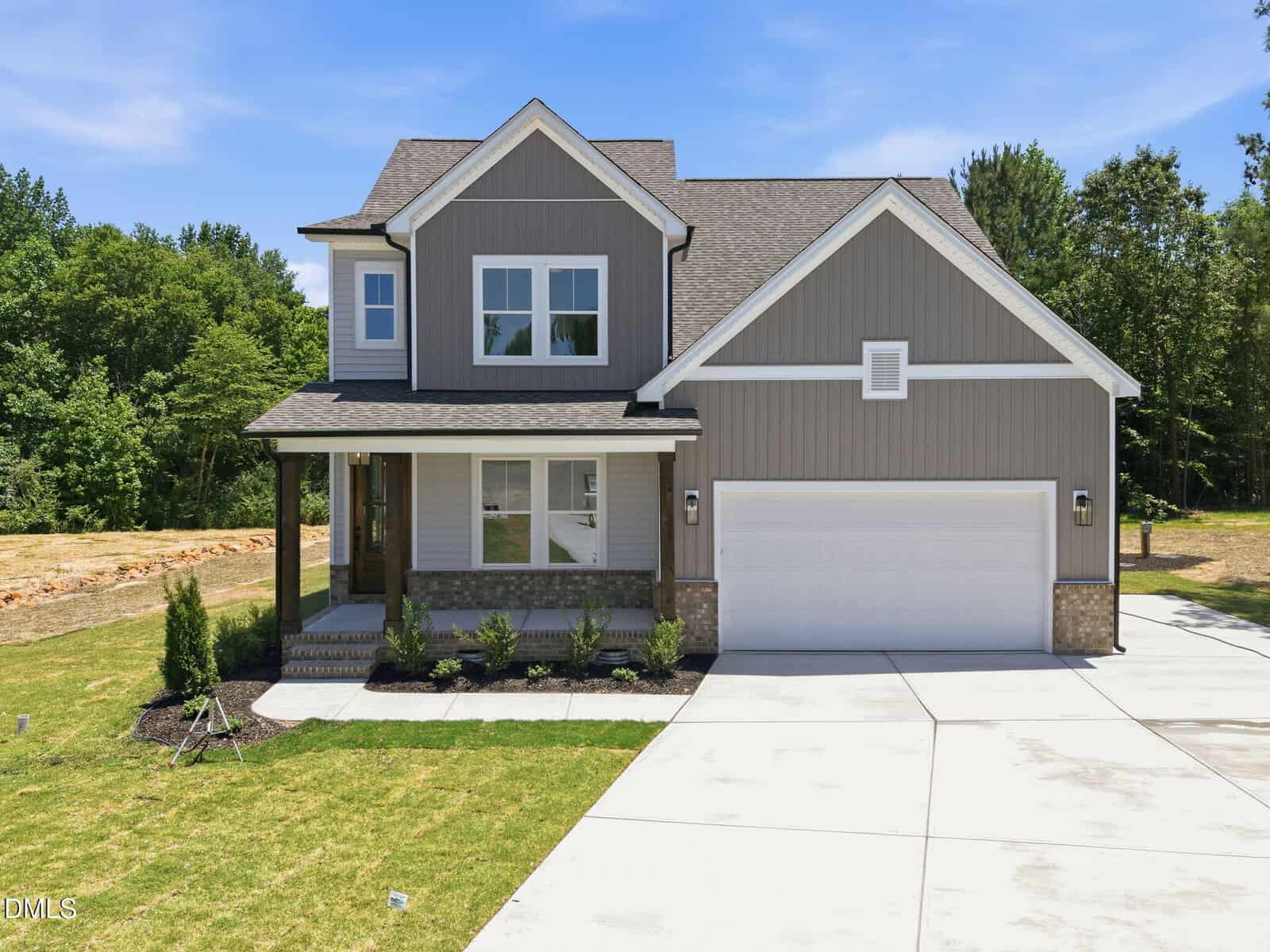 A modern two-story suburban house with gray siding, white trim, and a covered front porch. The home has a two-car garage and a neat front lawn with small shrubs, set against a backdrop of trees and blue sky.