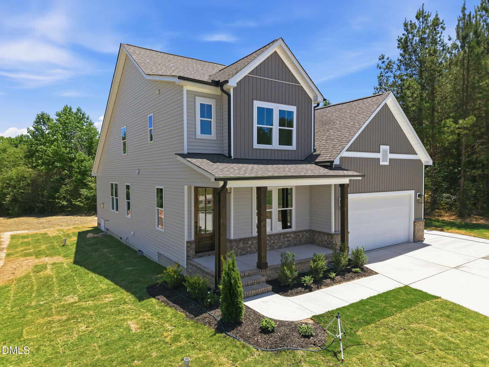A two-story modern house with gray siding, white trim, a covered front porch with wooden pillars, attached two-car garage, and landscaped front yard, set against a backdrop of trees under a blue sky.