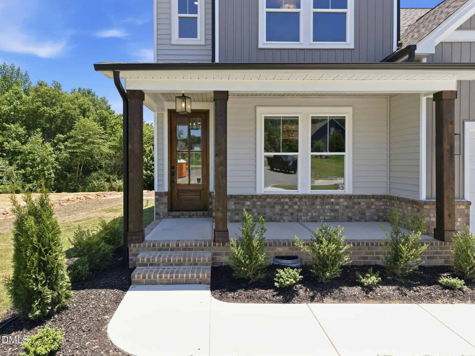 A modern house exterior with beige siding, brick accents, wooden columns, a covered front porch, and shrubs along the walkway. Large windows reflect the outdoors on a sunny day.