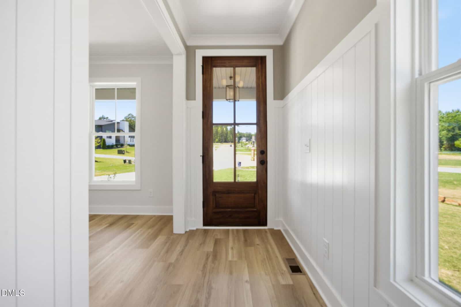 A bright entryway with white paneled walls, light wood floors, and a dark wooden front door with glass inset. Large windows let in natural light, showing views of a grassy neighborhood outside.