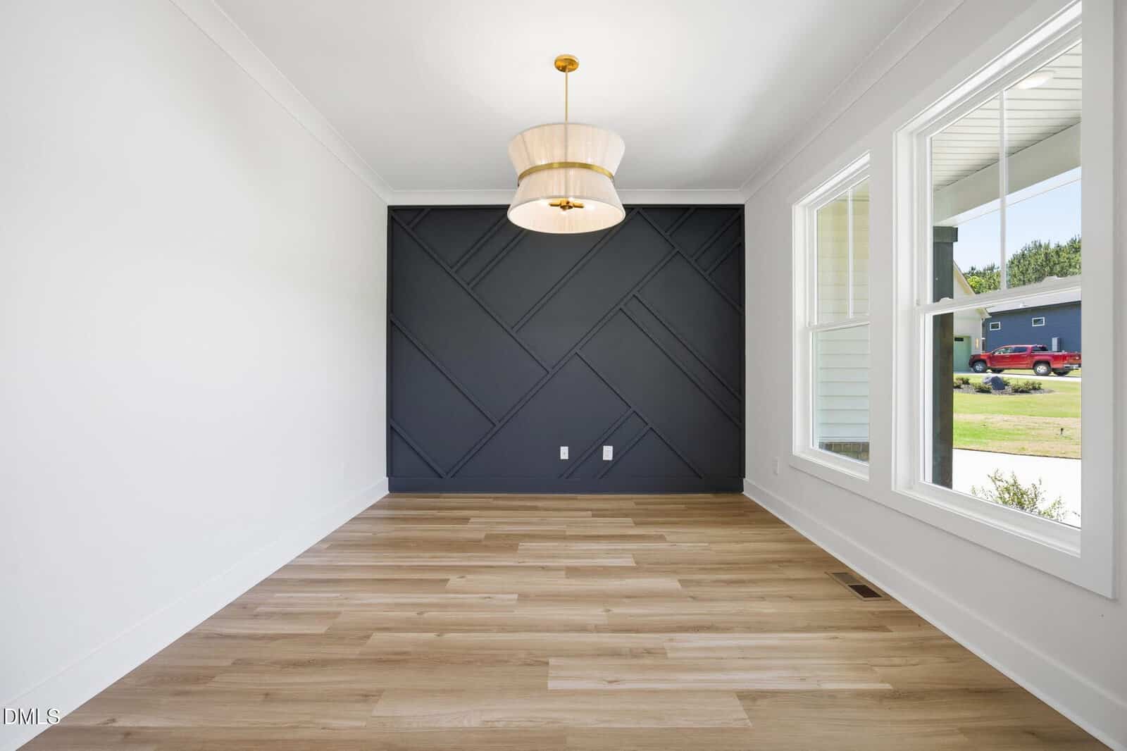 Modern, empty room with light wood floors, a geometric black accent wall, large windows letting in sunlight, and a gold pendant light fixture hanging from the white ceiling.
