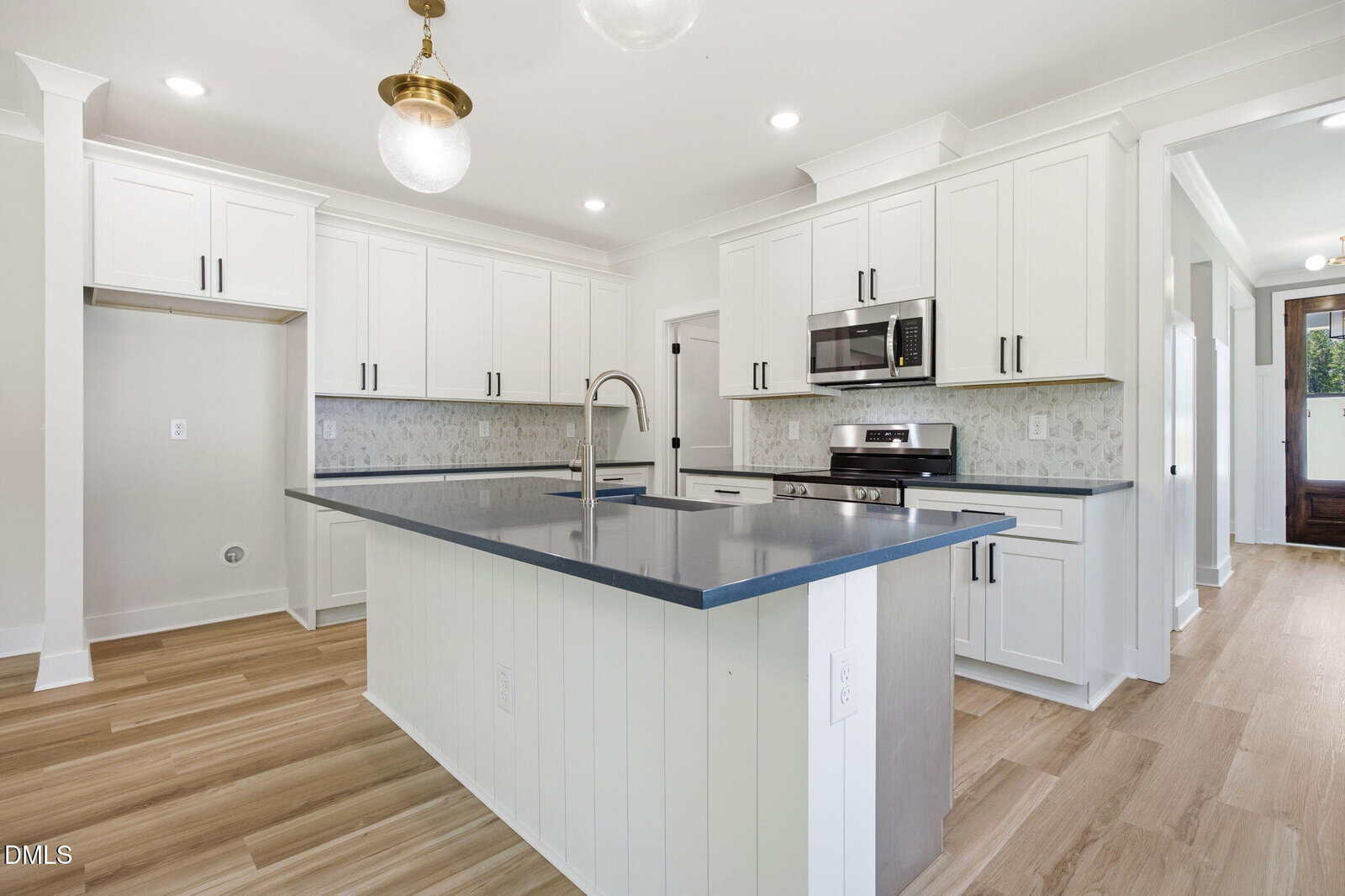 Modern kitchen with white cabinets, stainless steel appliances, a gray countertop island with built-in sink, light wood floors, and pendant lighting. There is space for a refrigerator and a built-in microwave above the stove.