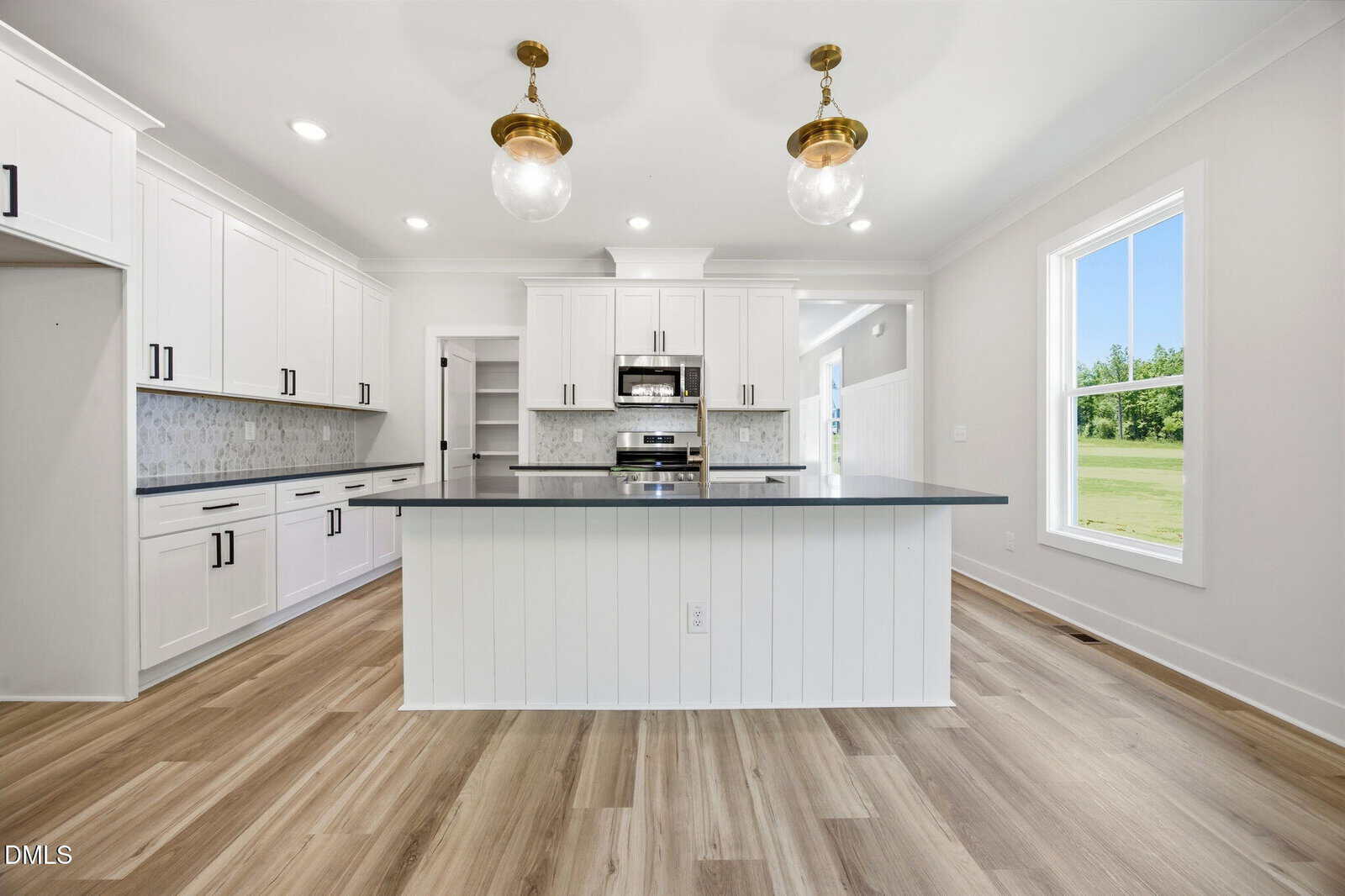 Modern white kitchen with a large island, gray countertop, wood floors, stainless steel appliances, glass pendant lights, and a window showing a green outdoor view.