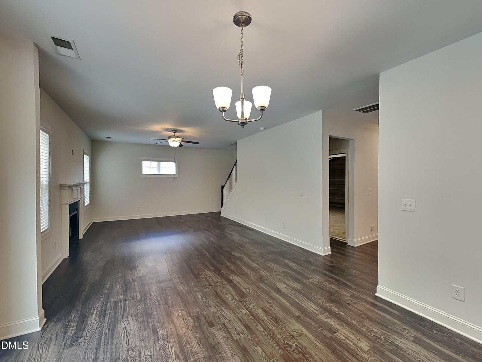 Spacious living room with dark wood floors, light walls, ceiling fans, chandelier, fireplace, windows, and an open doorway leading to a stairway and closet.