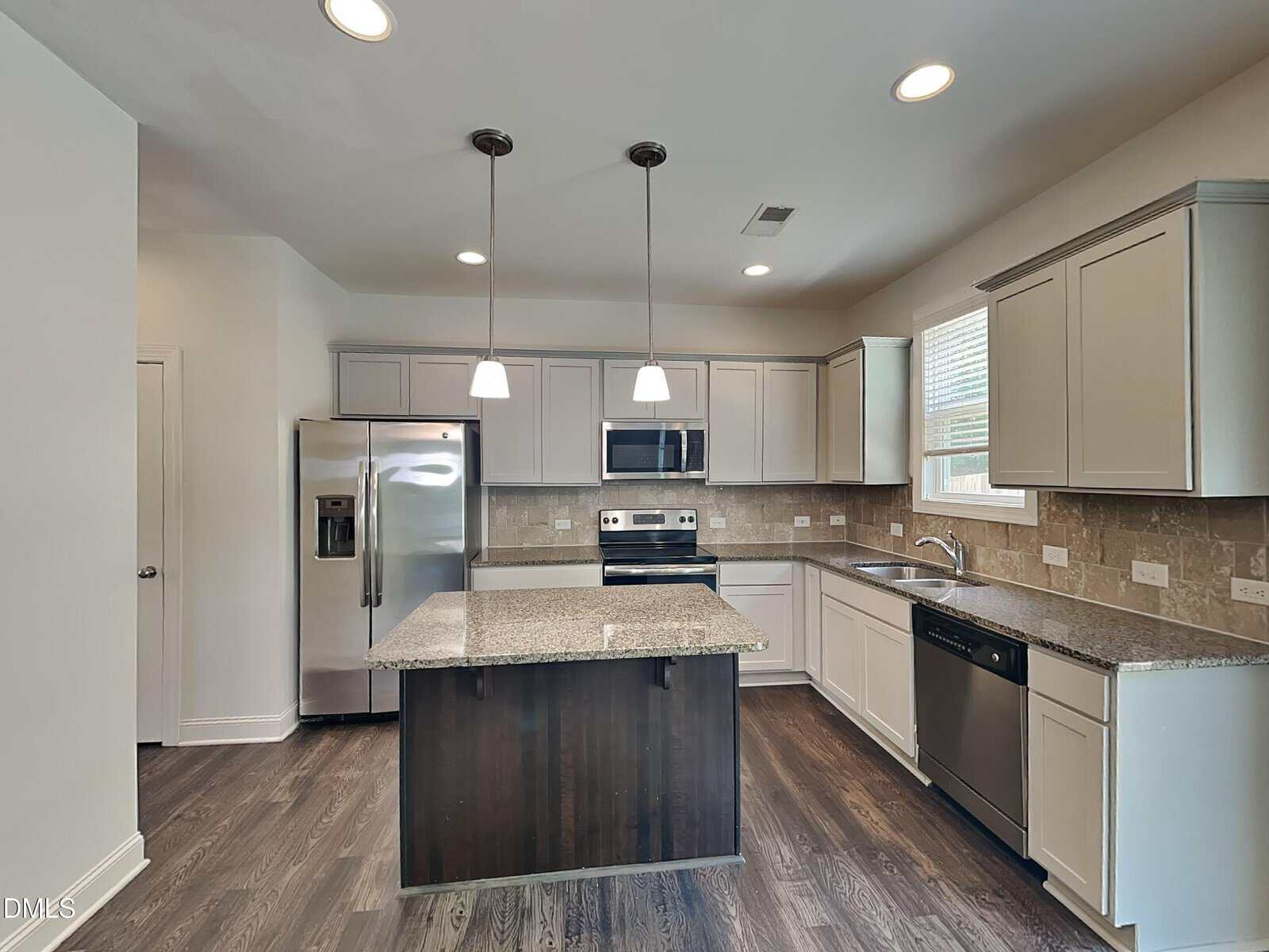 Modern kitchen with stainless steel appliances, an island with granite countertop, white cabinets, tile backsplash, two pendant lights, and dark wood flooring. Sunlight streams in through a window above the sink.