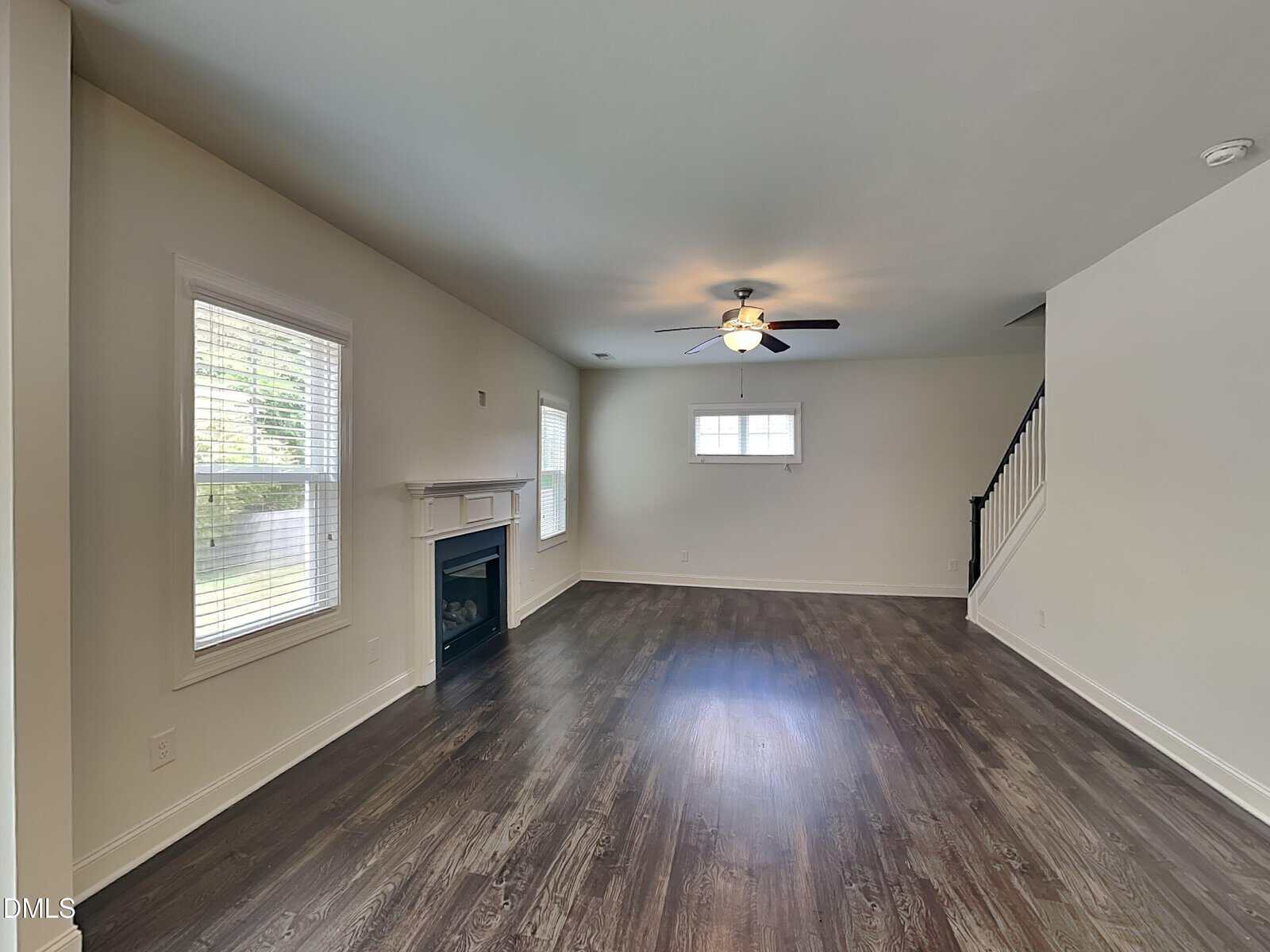 Spacious living room with dark wood flooring, white walls, a ceiling fan with light, windows allowing natural light, a fireplace, and a staircase leading to the upper floor.