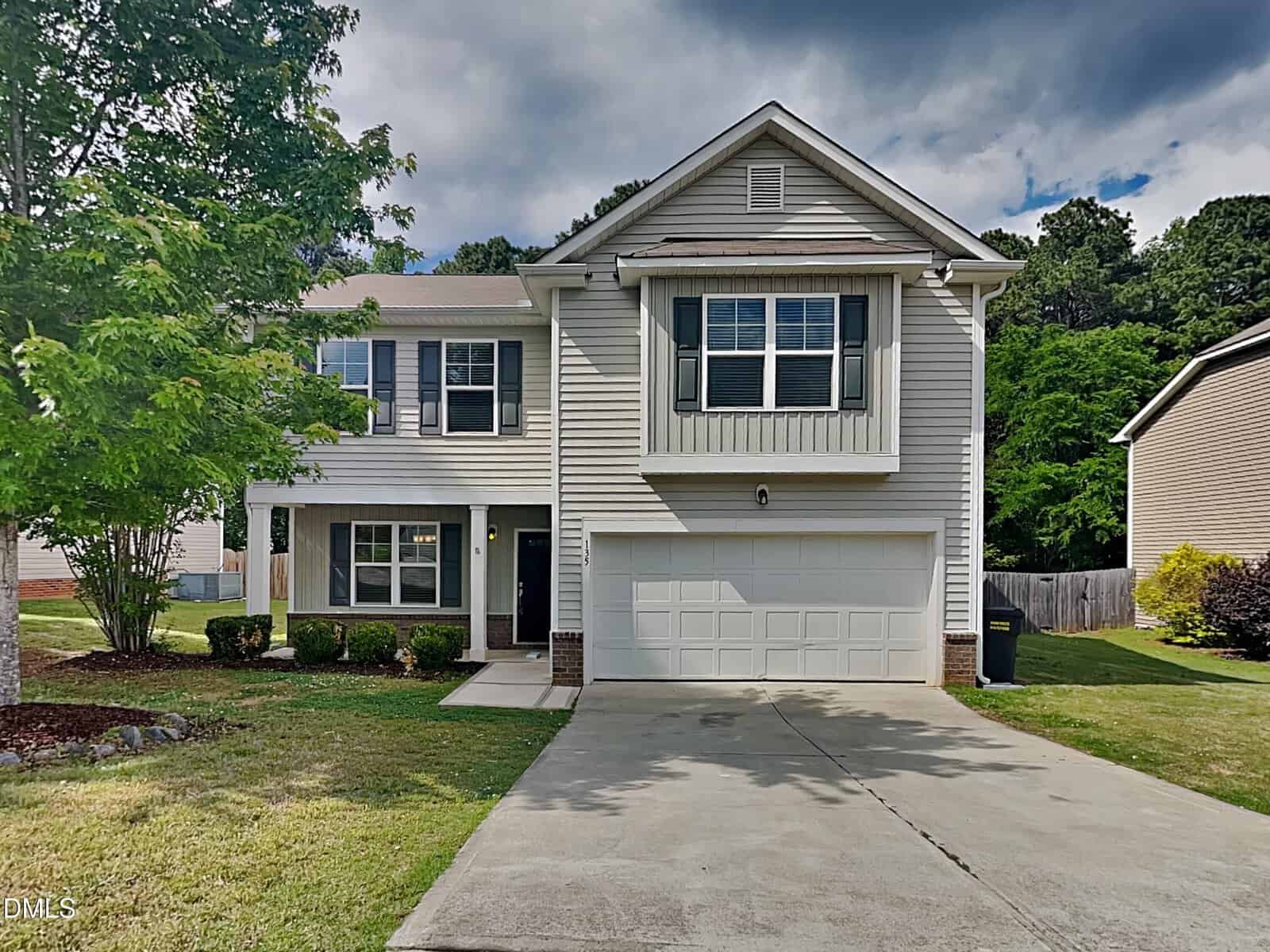Two-story suburban house with light gray siding, white trim, and a two-car garage. A tree and green lawn are in front, with a concrete driveway leading to the garage. Shrubs border the walkway to the front door.