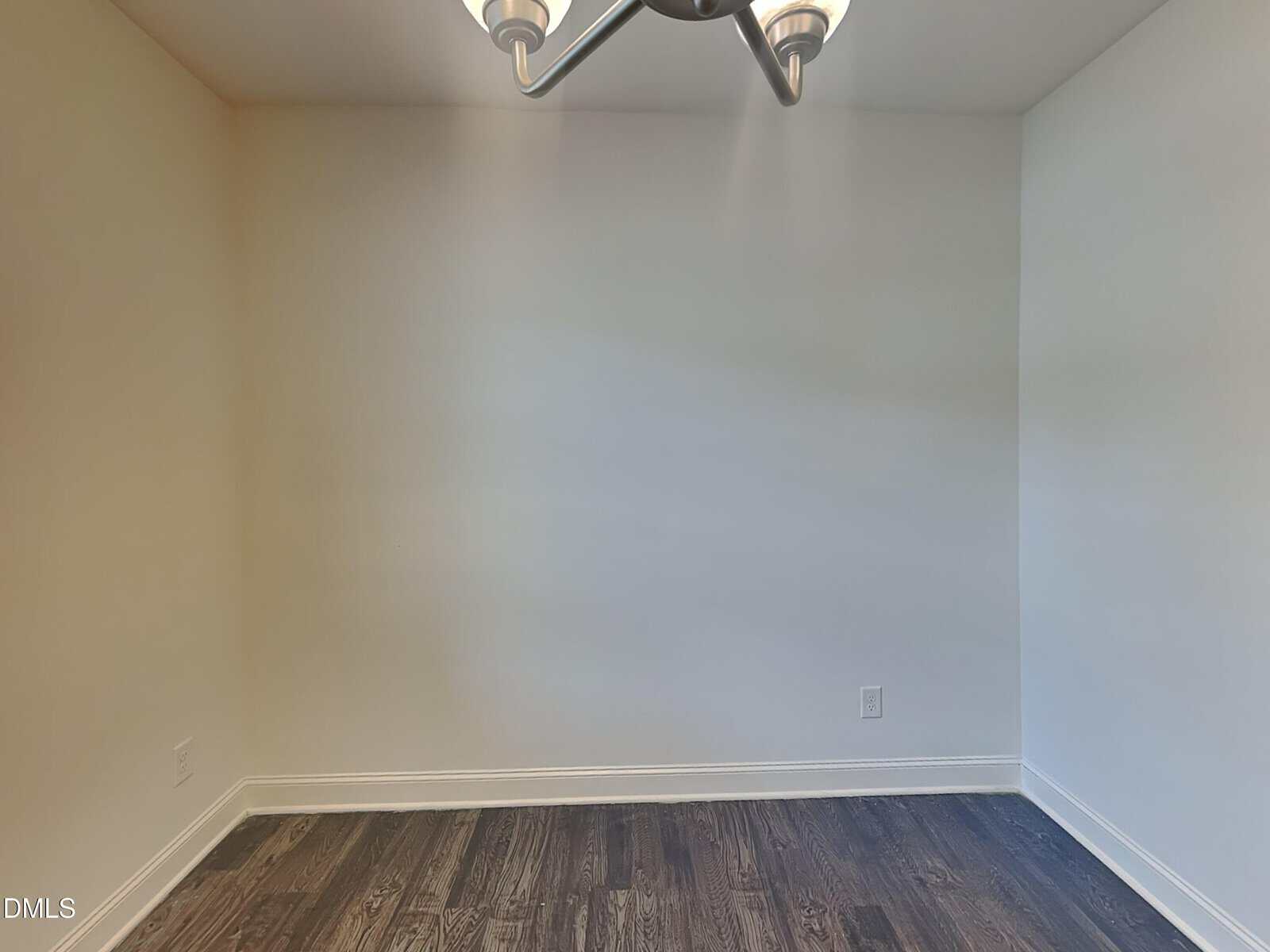Empty room with white walls, dark hardwood flooring, and a modern ceiling light fixture, photographed from one corner. There are no windows or furniture visible in the image.