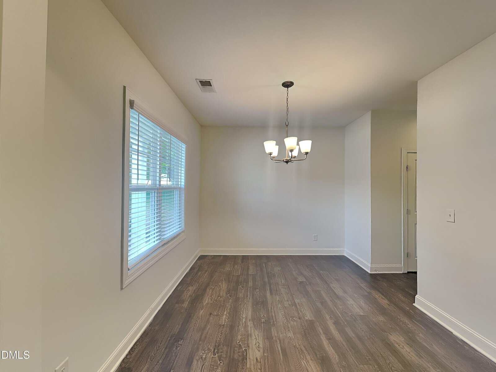 A bright dining area with light-colored walls, a large window with blinds on the left, a modern chandelier hanging from the ceiling, and dark wood flooring. A small doorway is visible on the right.