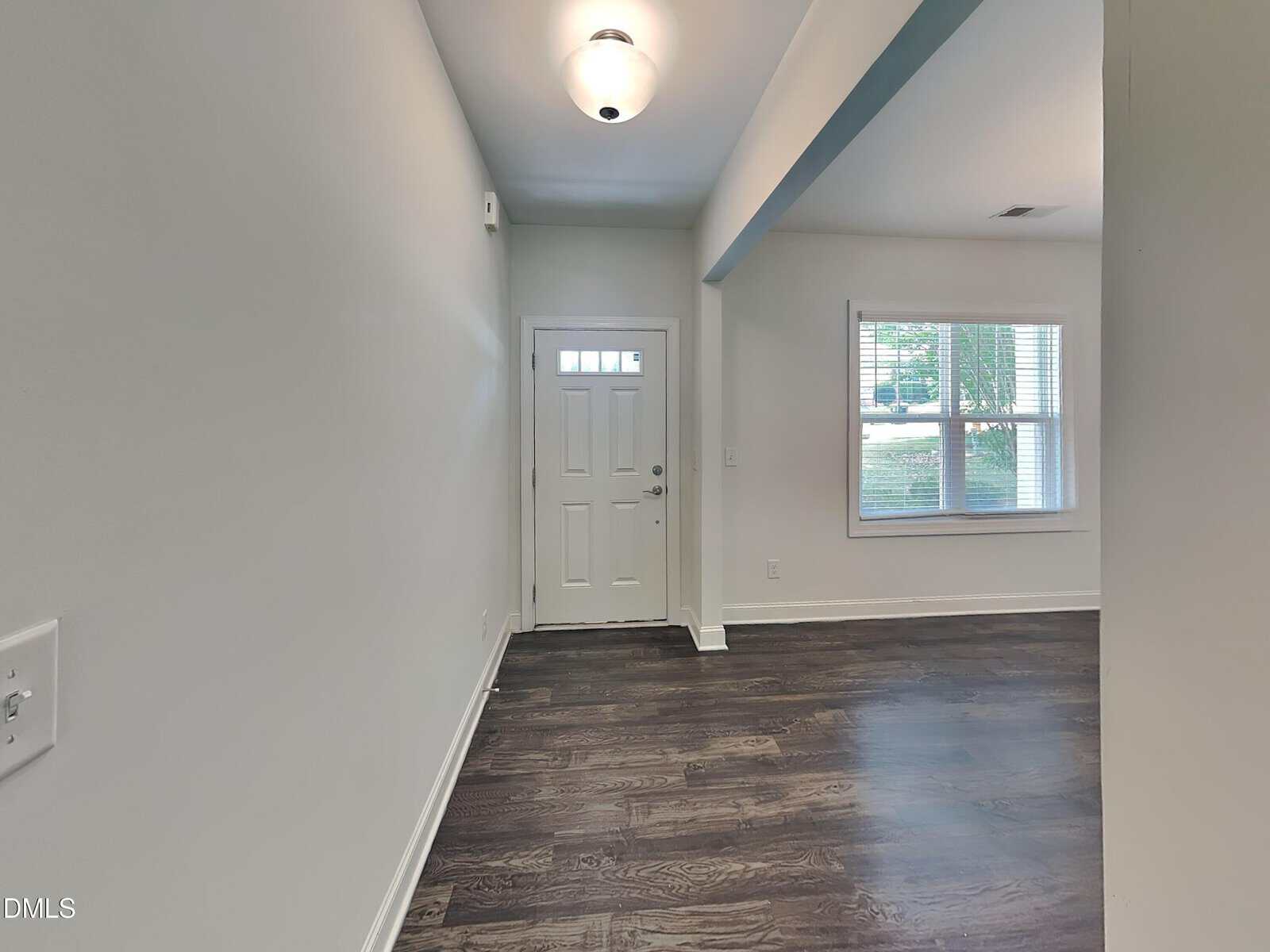 Entryway with a white front door, adjacent window, and dark wood floors. Light gray walls and a ceiling light fixture are visible, along with natural light coming through the window.