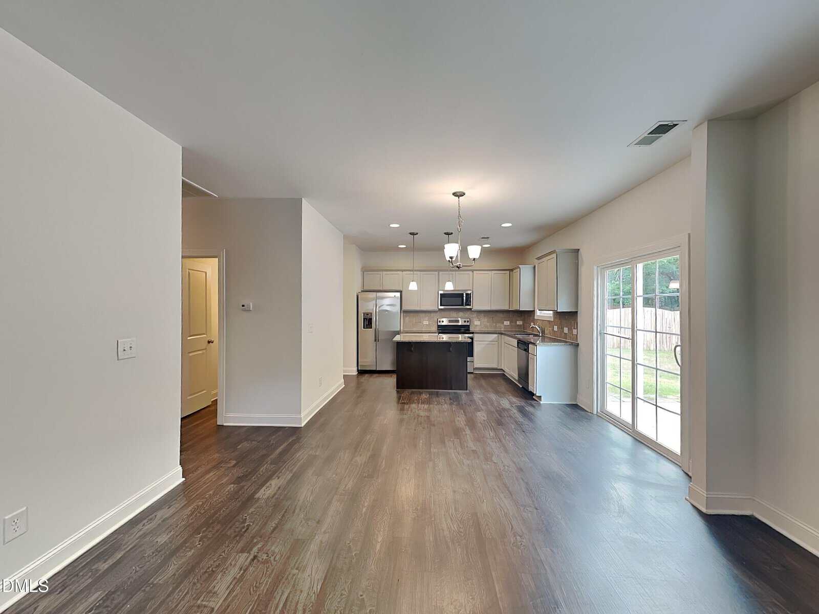 Open-concept kitchen and dining area with dark wood floors, stainless steel appliances, white cabinets, and sliding glass doors leading to a fenced backyard. The space is well-lit with natural and ceiling lights.