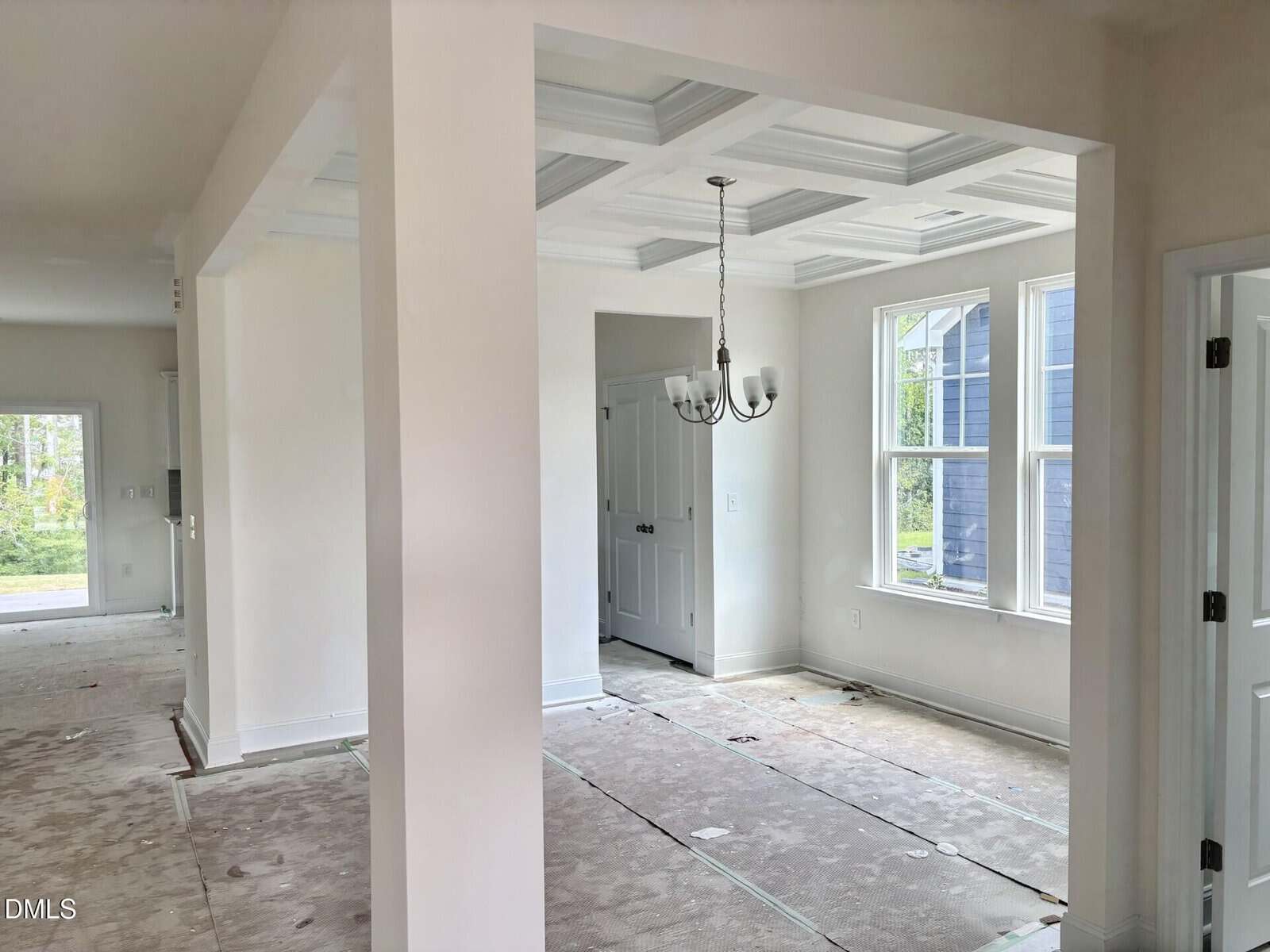 Empty, partially finished room with white walls, coffered ceiling, and chandelier. Protective coverings are on the floor. Natural light enters through large windows. A door and hallway are visible in the background.